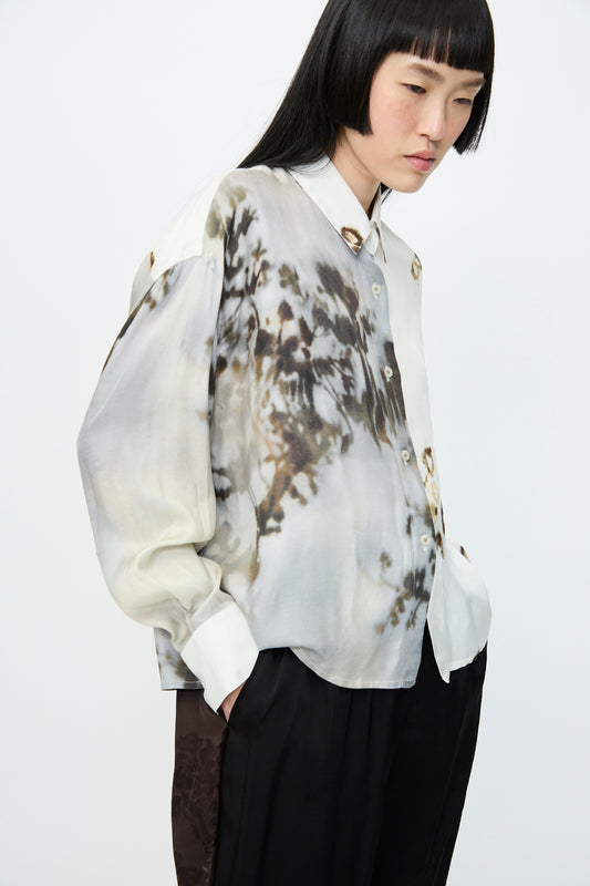 Person with black hair wears a Cordera Printed Classic Shirt in Bicolor, oversized and patterned with printed viscose, paired with dark pants, hands casually in pockets against a plain background.