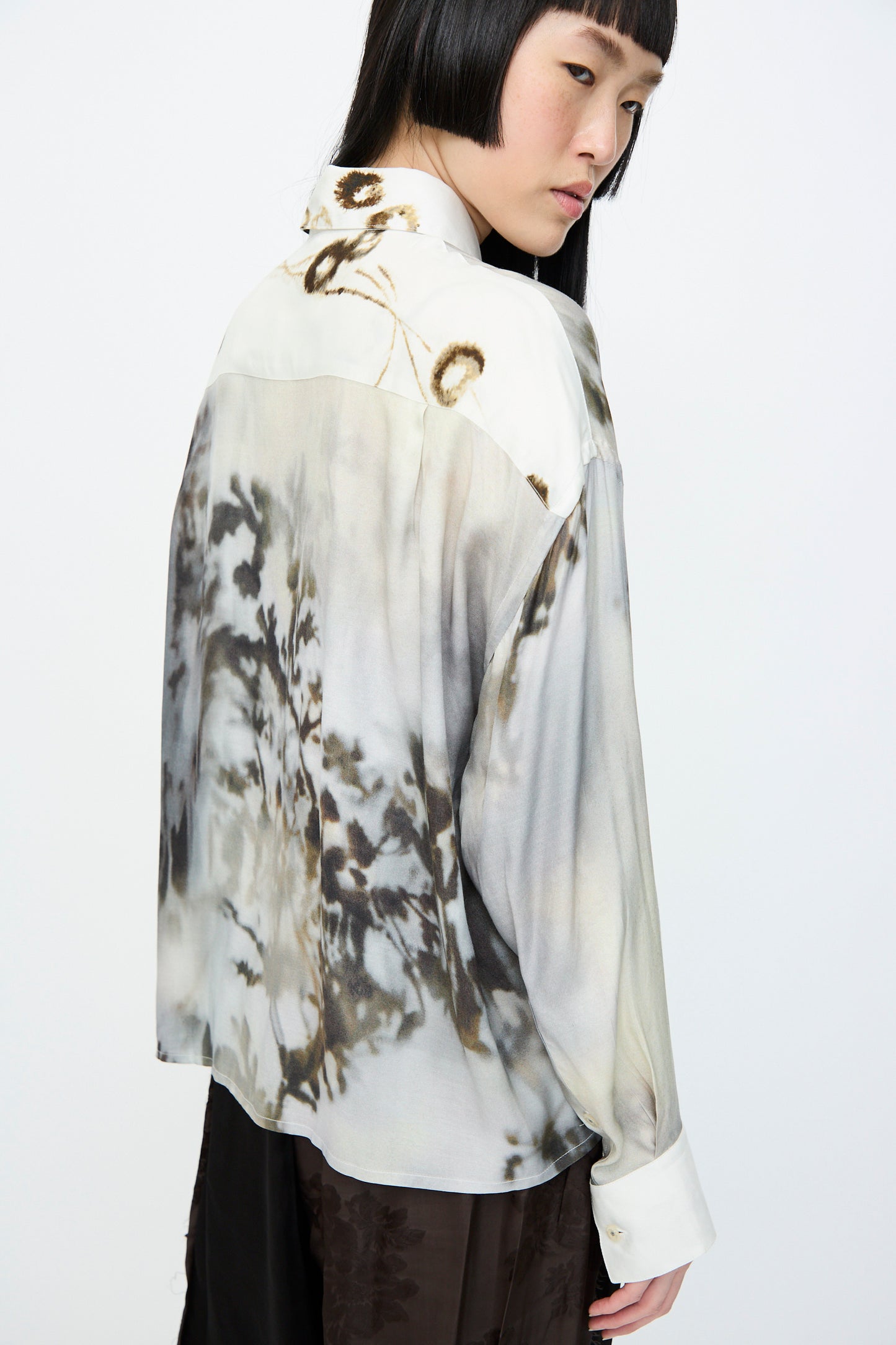 Person in an oversized, long-sleeve Printed Classic Shirt in Bicolor by Cordera and dark pants, looking away against a plain background.