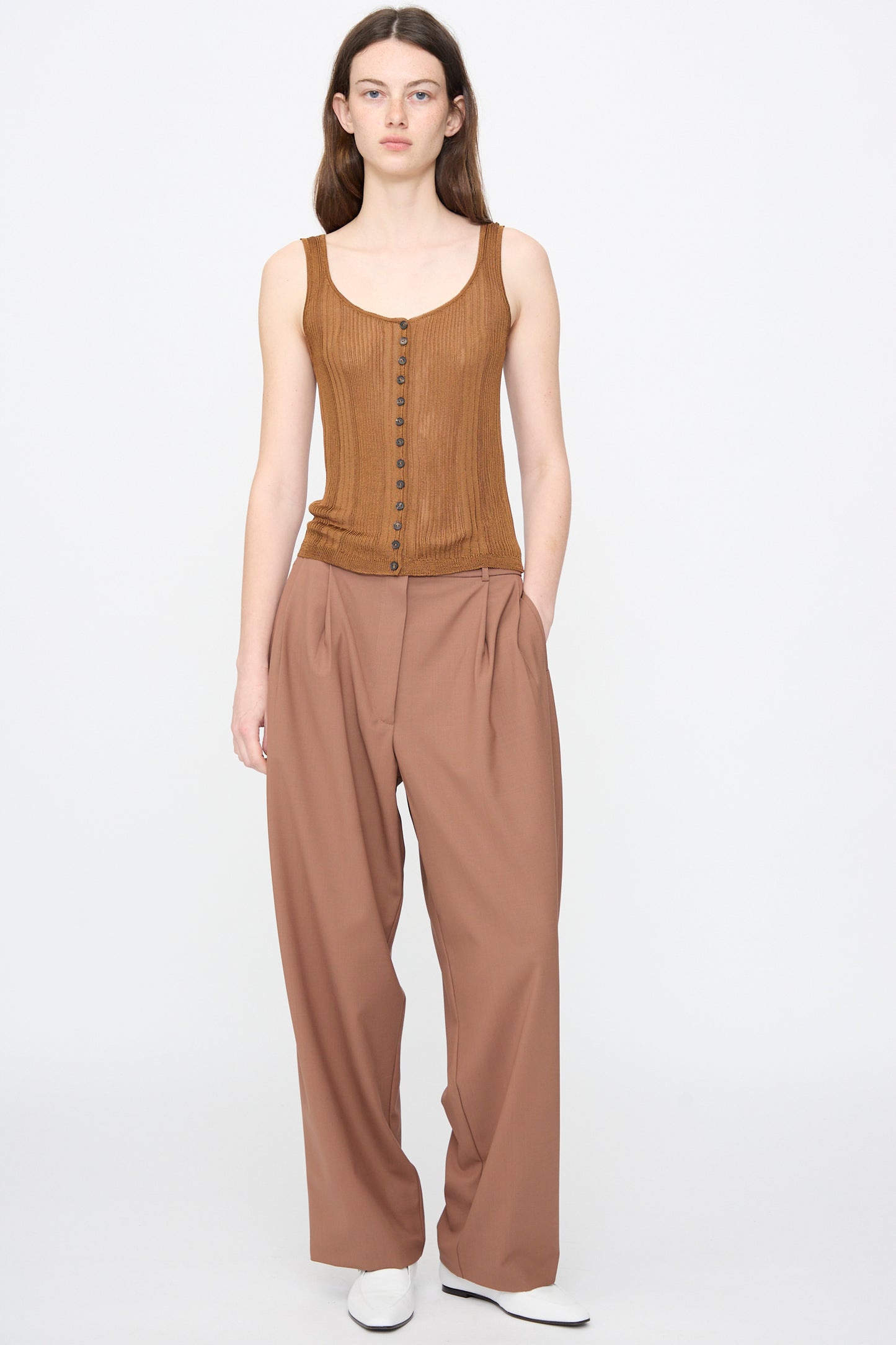 A person stands facing forward against a plain white background, wearing Cordera's Ribbed Tank Top in Tobacco, paired with wide-legged brown trousers and white shoes.