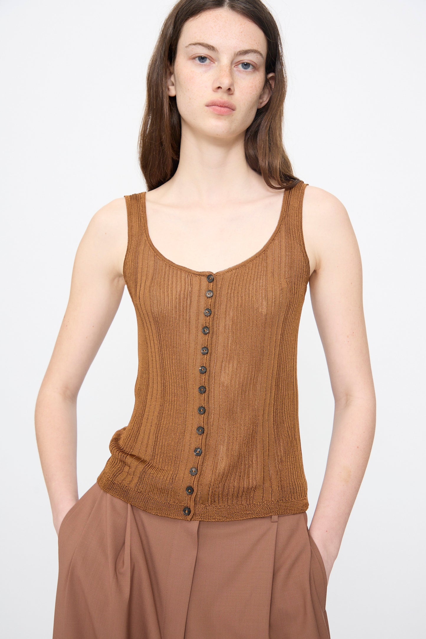 A person with long brown hair wears the Cordera Ribbed Tank Top in Tobacco and loose brown pants, standing against a plain light background.