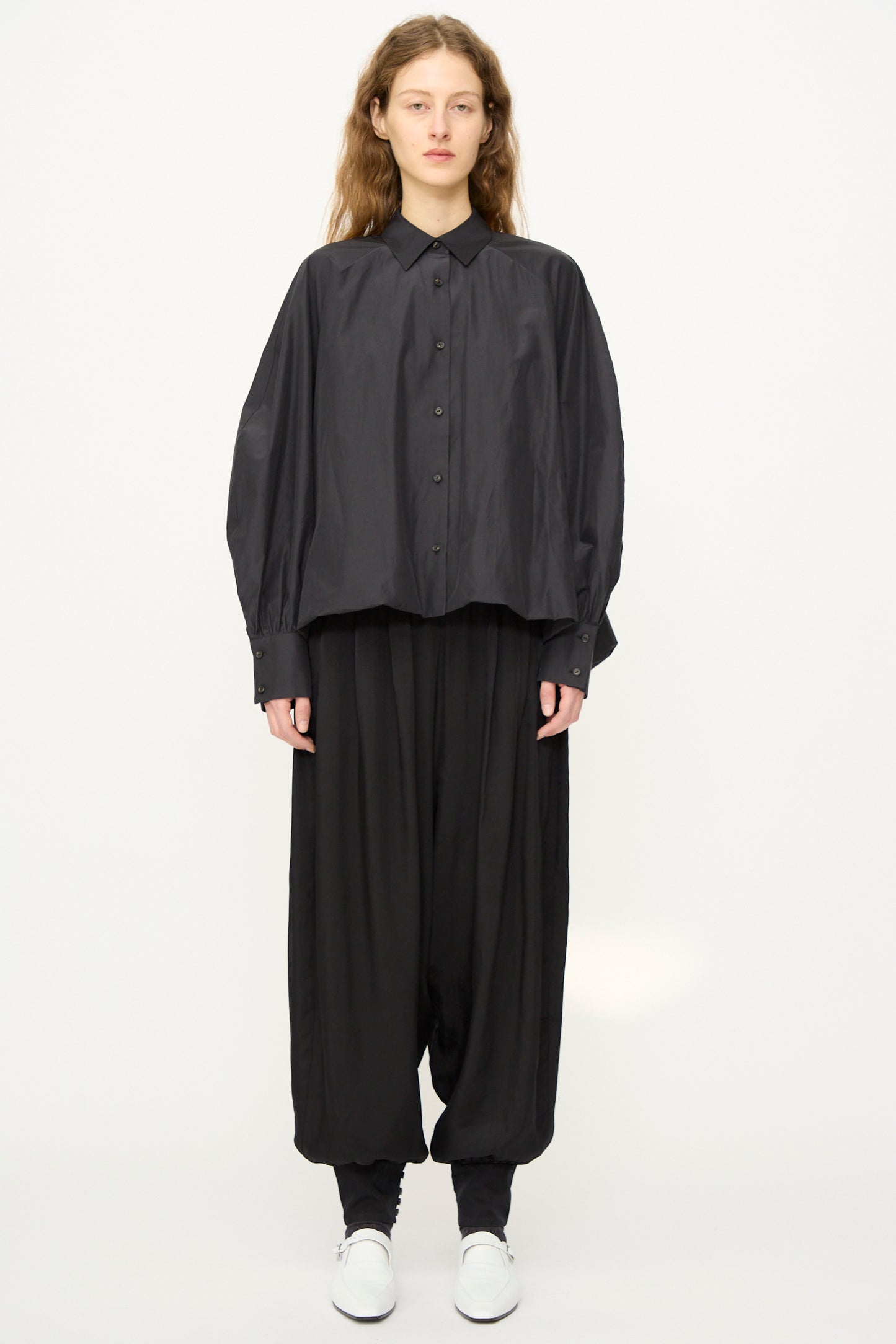 Person stands against a plain background wearing Cordera's Sarouel Trouser in Black, a loose-fitting black button-up shirt, black leggings, low-rise balloon-shaped trousers, and white shoes.