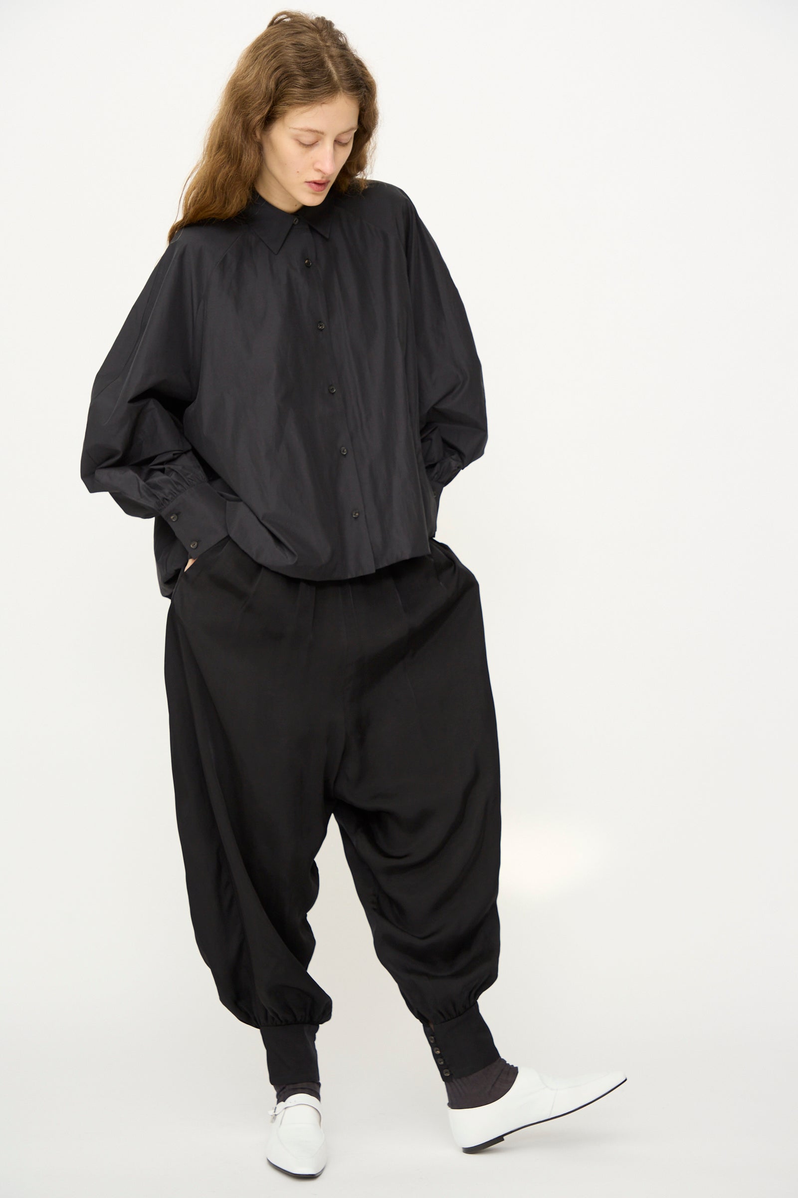 A person stands against a plain background wearing Cordera’s Sarouel Trouser in Black, paired with a loose black button-up shirt, white shoes, and black socks.