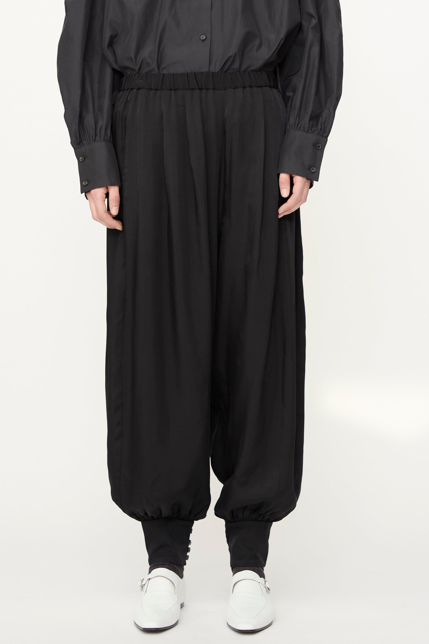 A model wears the Cordera Sarouel Trouser in Black—loose, balloon-shaped pants with elastic cuffs—paired with a black button-up shirt and white shoes against a plain light background.