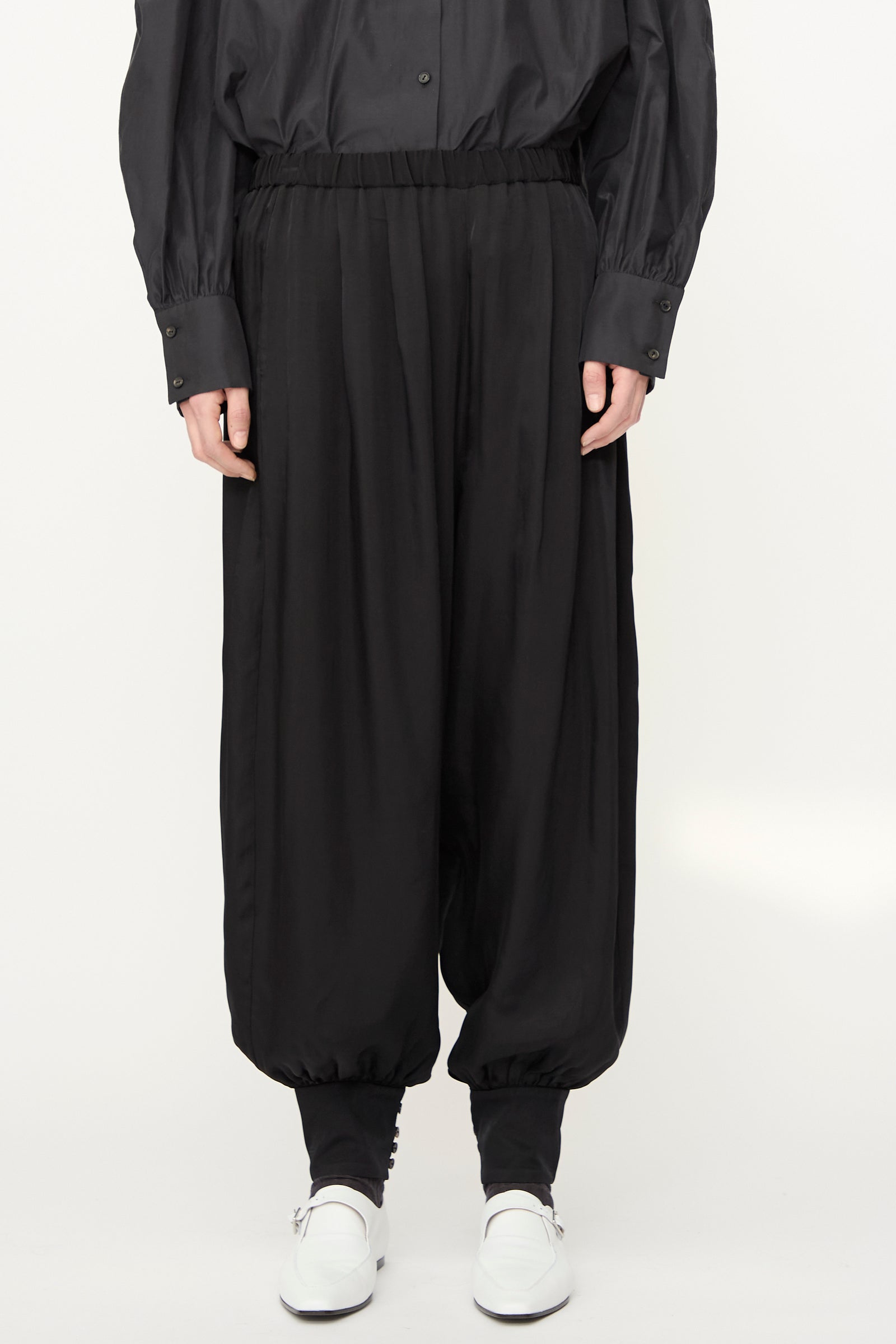 A model wears the Cordera Sarouel Trouser in Black—loose, balloon-shaped pants with elastic cuffs—paired with a black button-up shirt and white shoes against a plain light background.