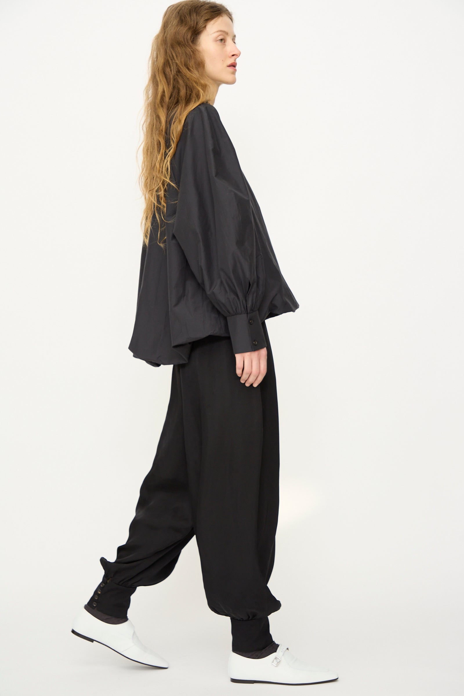 Model wearing Cordera's Sarouel Trouser in Black—a loose, balloon-shaped, low-rise style—paired with a relaxed black shirt and white shoes, walking in profile against a plain white background.