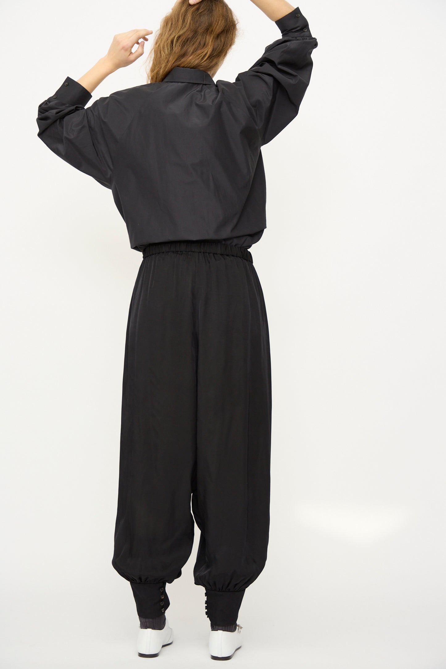 A person stands with back to camera, holding up their hair, wearing a black long-sleeve shirt and Cordera's Sarouel Trouser in Black with elastic cuffs and loose fit, set against a plain white background.