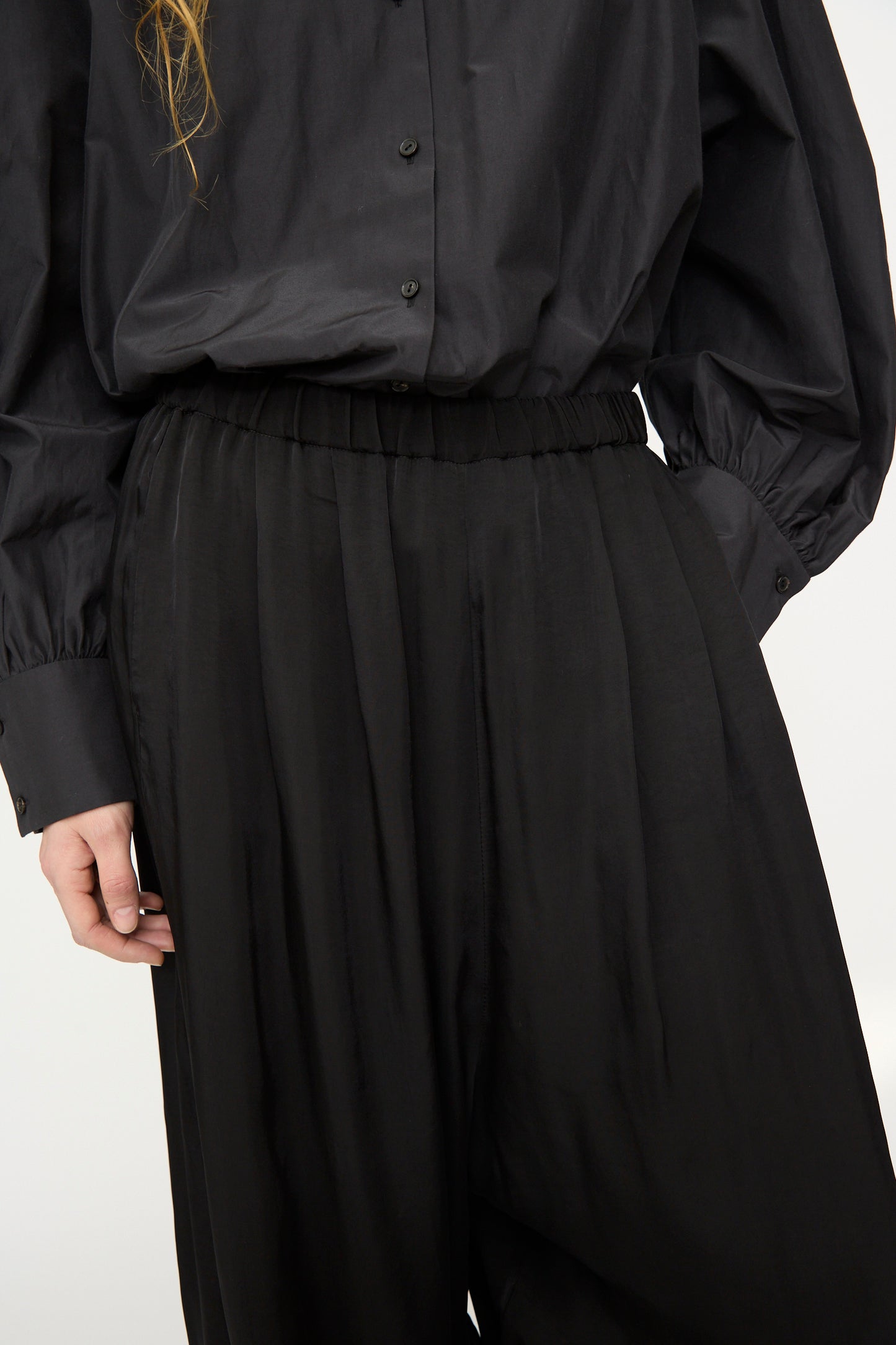 A person wears Cordera’s Sarouel Trouser in Black with a loose black button-up shirt, showcasing wide, balloon-shaped legs and one hand visible at their side.