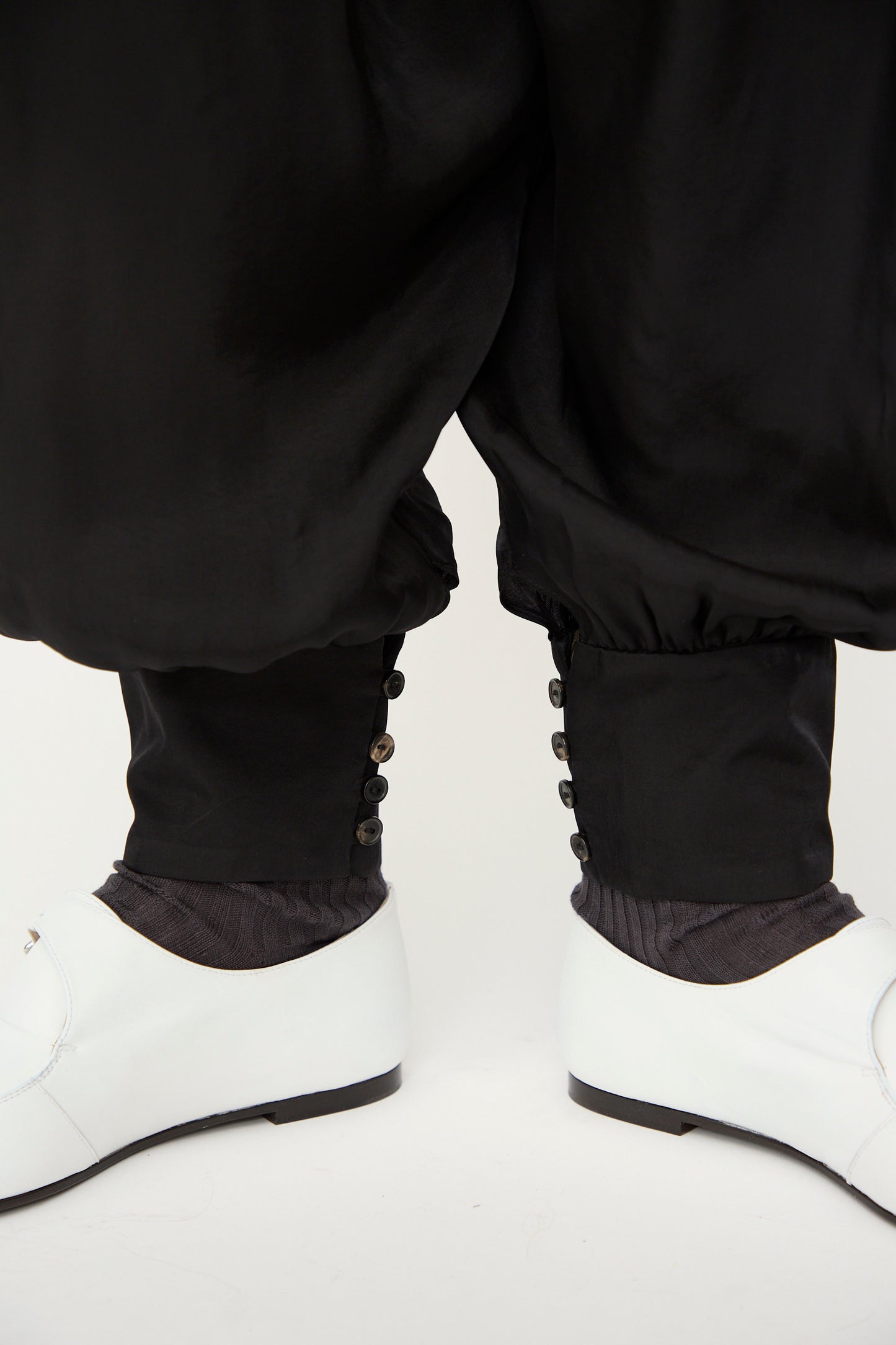 Close-up of a person wearing Cordera's Sarouel Trouser in Black, featuring balloon-shaped legs and buttoned cuffs, styled with black socks and white dress shoes against a plain background.
