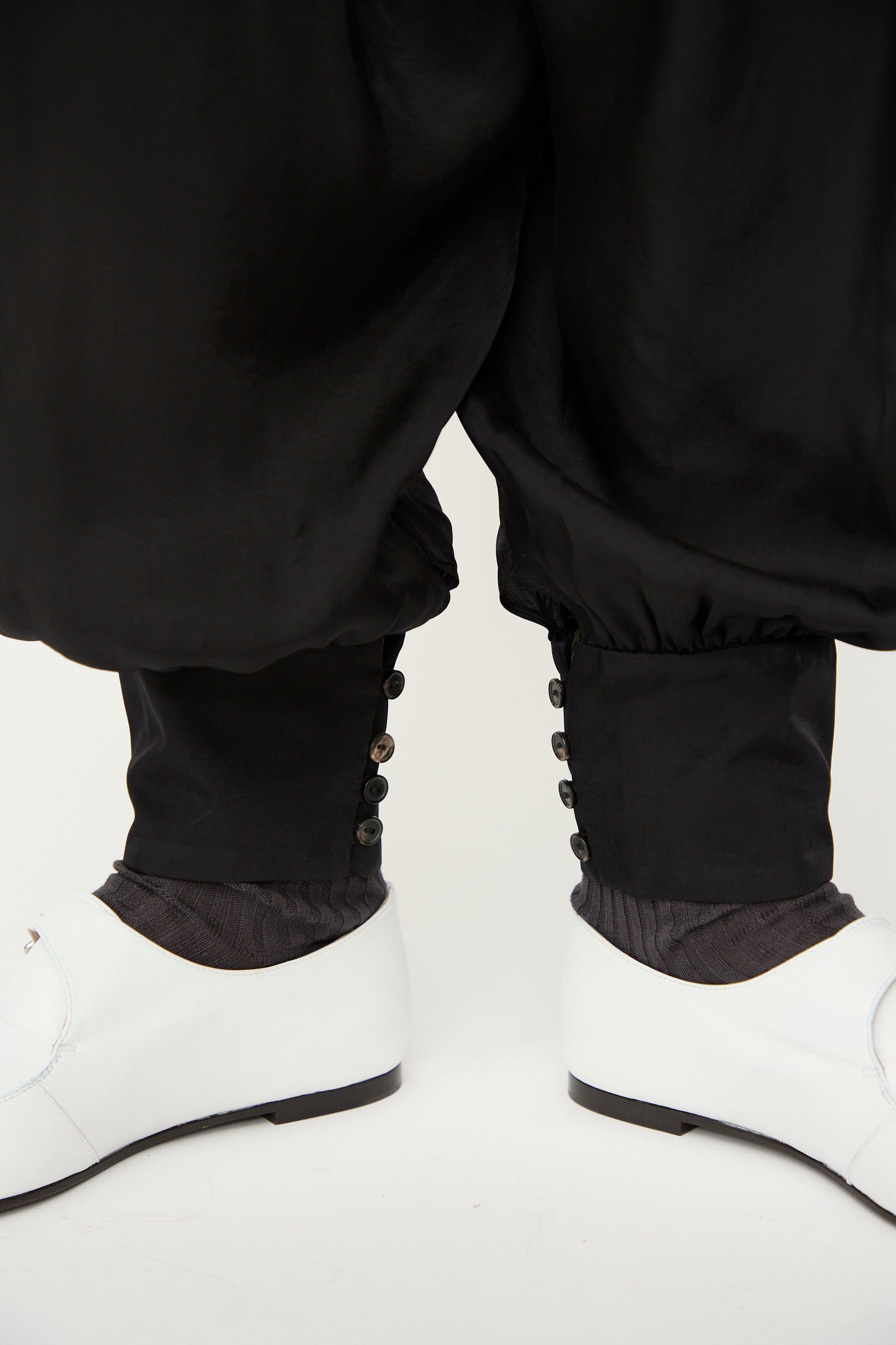Close-up of a person wearing Cordera's Sarouel Trouser in Black, featuring balloon-shaped legs and buttoned cuffs, styled with black socks and white dress shoes against a plain background.