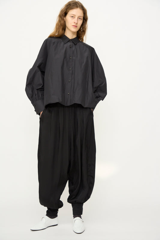 Person stands against a plain background wearing Cordera's Sarouel Trouser in Black, a loose black button-up shirt, black socks, and white shoes, with hands casually tucked in pockets.