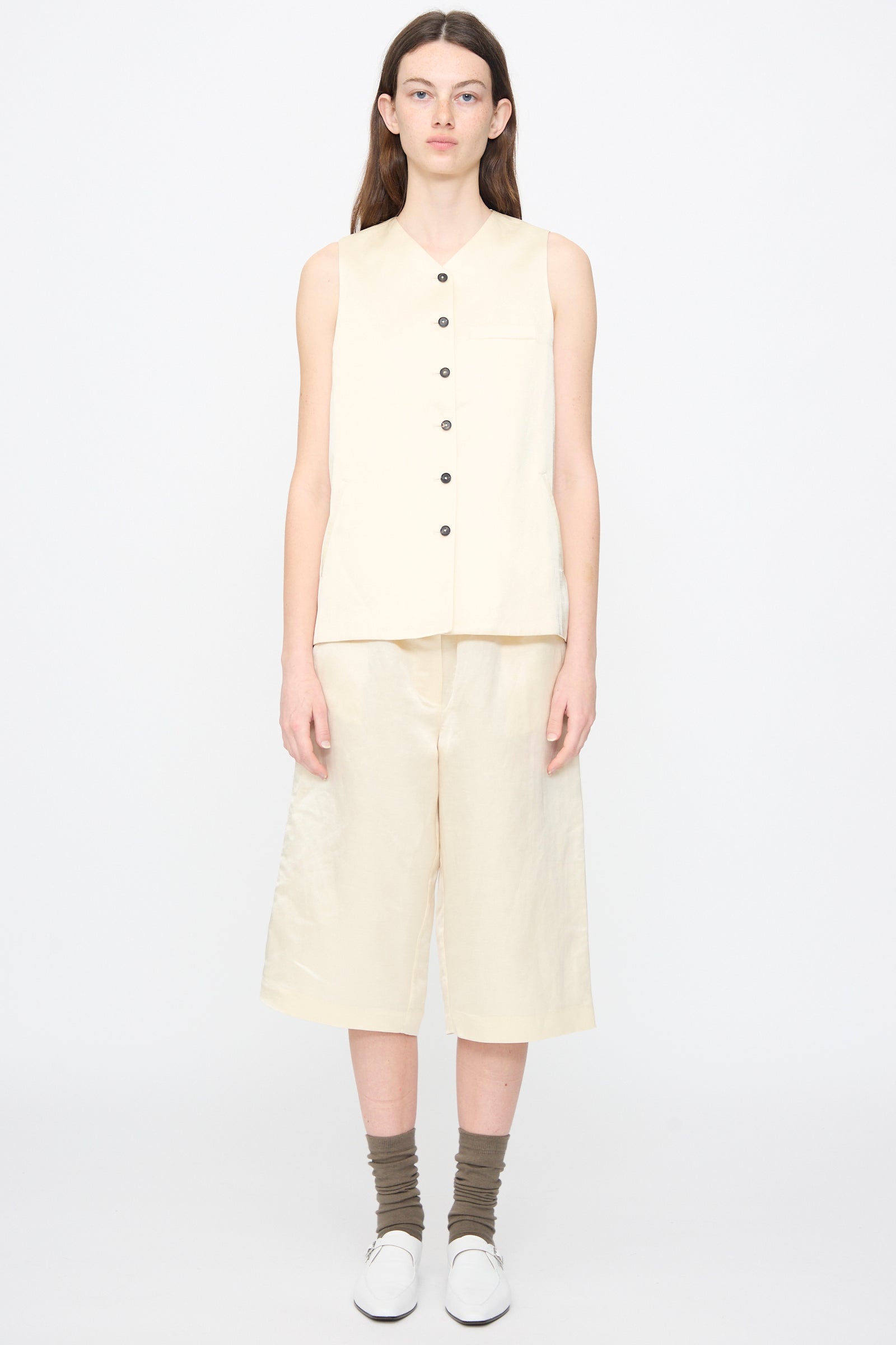 A person faces forward against a plain background, wearing a sleeveless cream button-up top, Cordera Satin Capri Pants in Off White, brown socks, and white shoes.