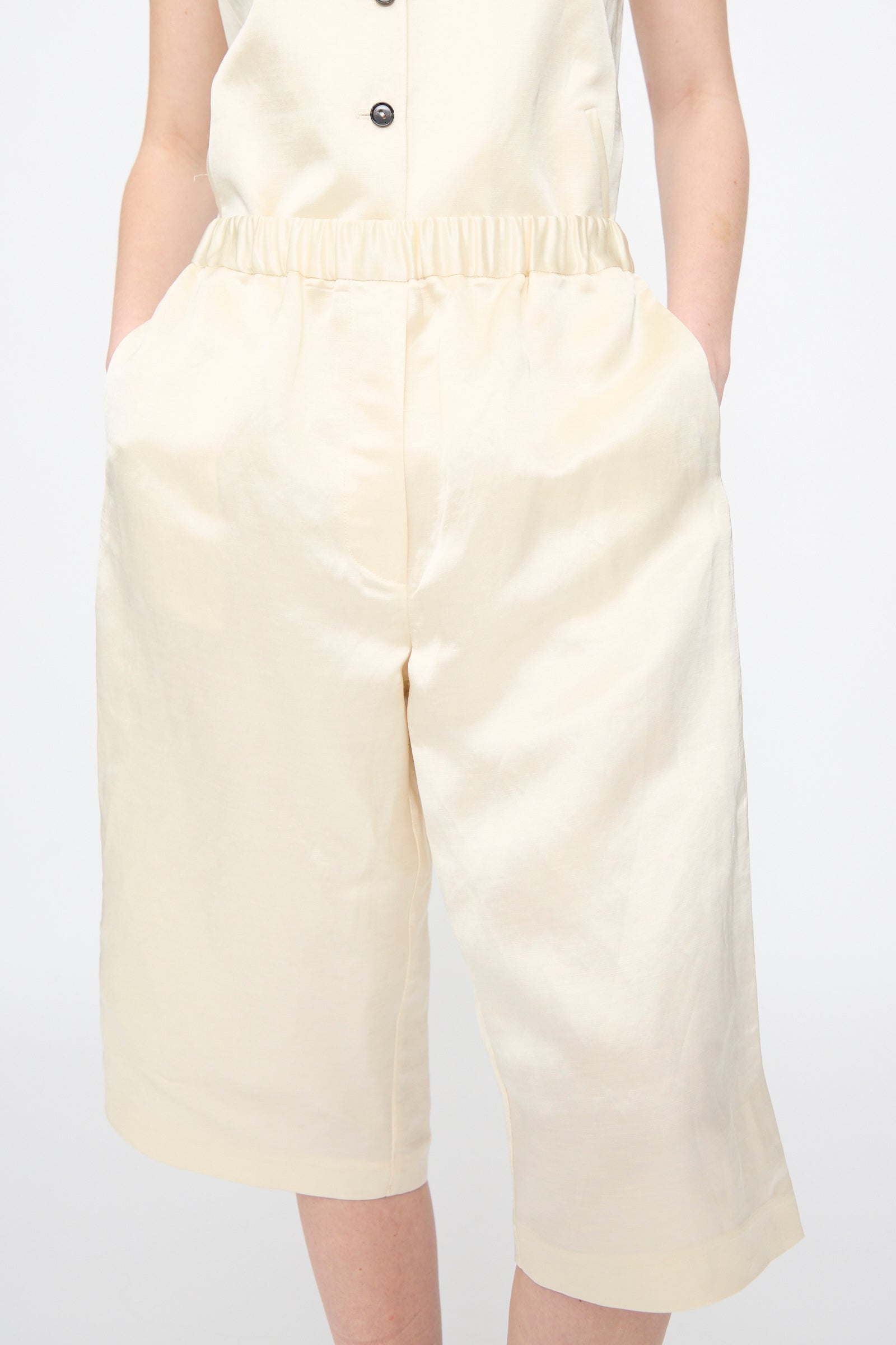 A person wearing Cordera’s Satin Capri Pants in Off White, featuring a relaxed fit and wide-leg, knee-length silhouette, standing with hands in their pockets.