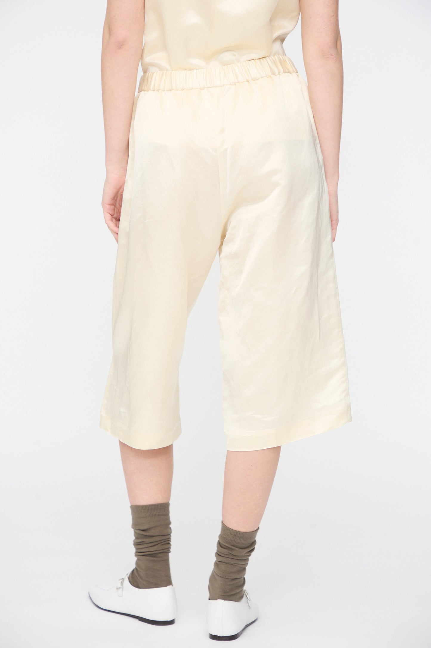 A person wears Cordera Satin Capri Pants in Off White with a matching top, brown socks, and white shoes, standing against a plain white background.