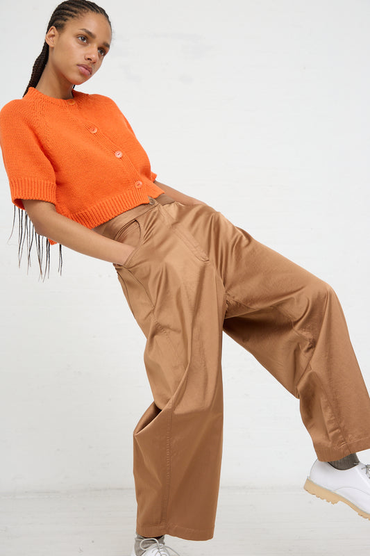 A woman posing in an orange cropped cardigan and high-rise, relaxed fit pants with one hand in her pocket, Cordera's Satin Curved Pant in Camel.