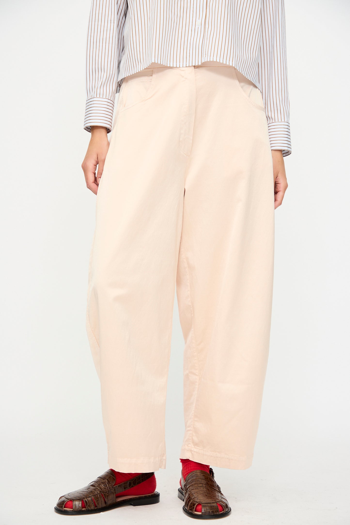 A person wearing Cordera’s Satin Curved Pant in Peach with an elastic waist, a striped long-sleeve shirt, red socks, and brown sandals stands against a plain white background.