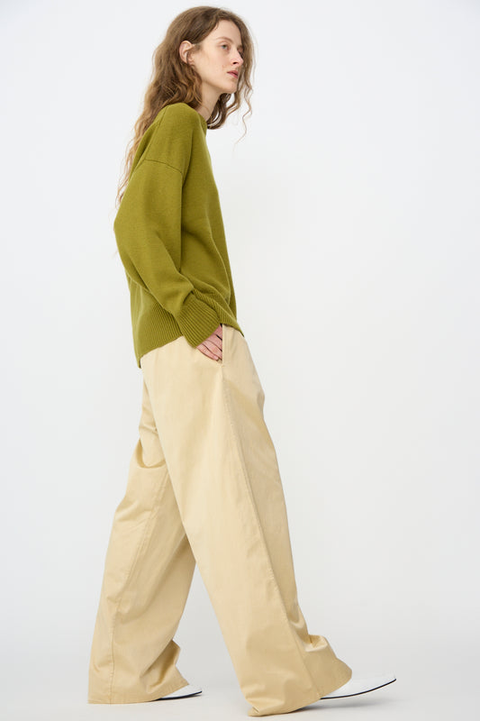 A person with long hair stands sideways against a white background, wearing an olive green sweater and Cordera's Satin Drawstring Pant in Champagne.