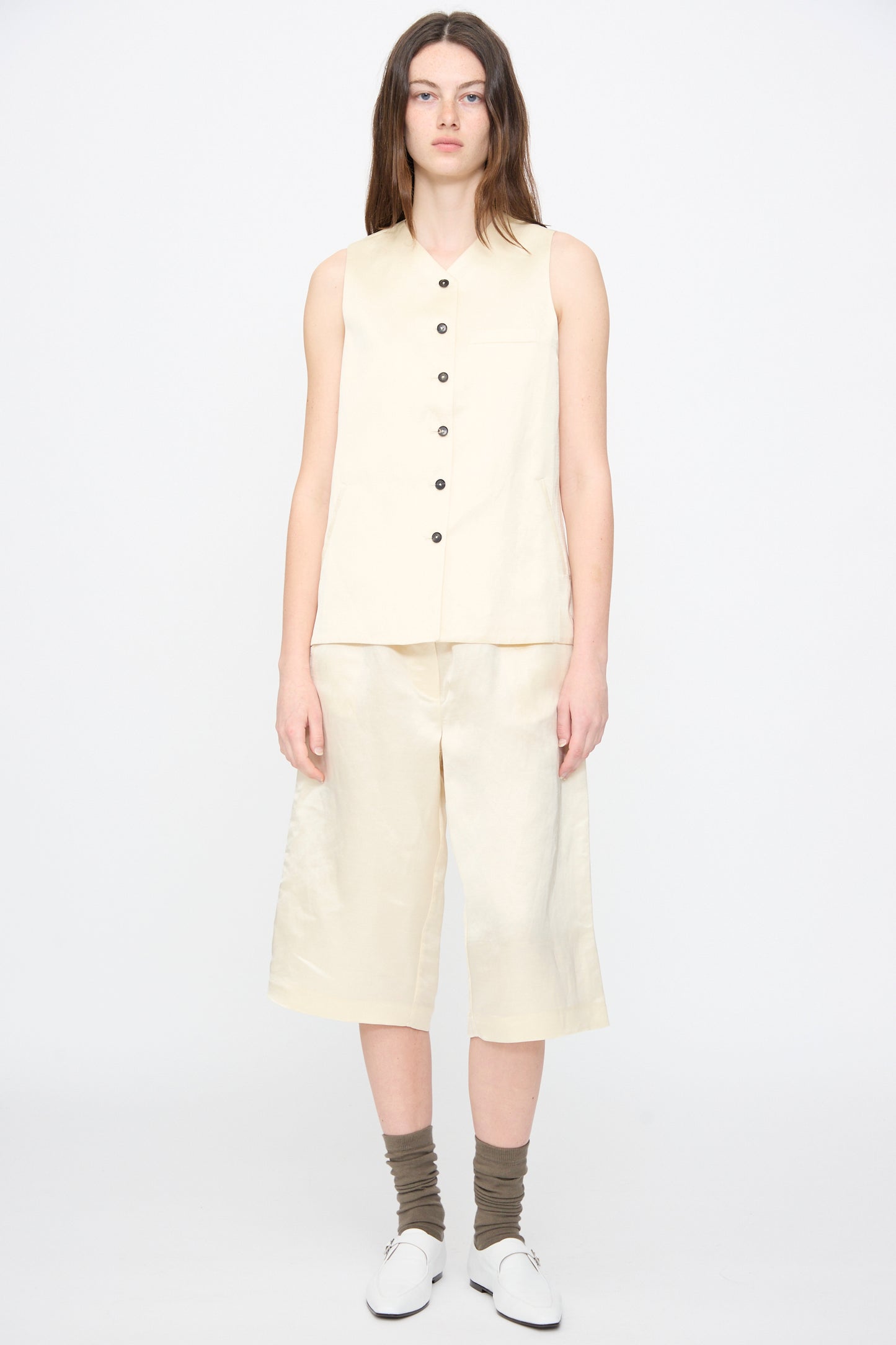 A person stands facing forward wearing the Satin Tailored Vest in Marfil by Cordera, matching knee-length shorts, gray socks, and white shoes against a plain light background.
