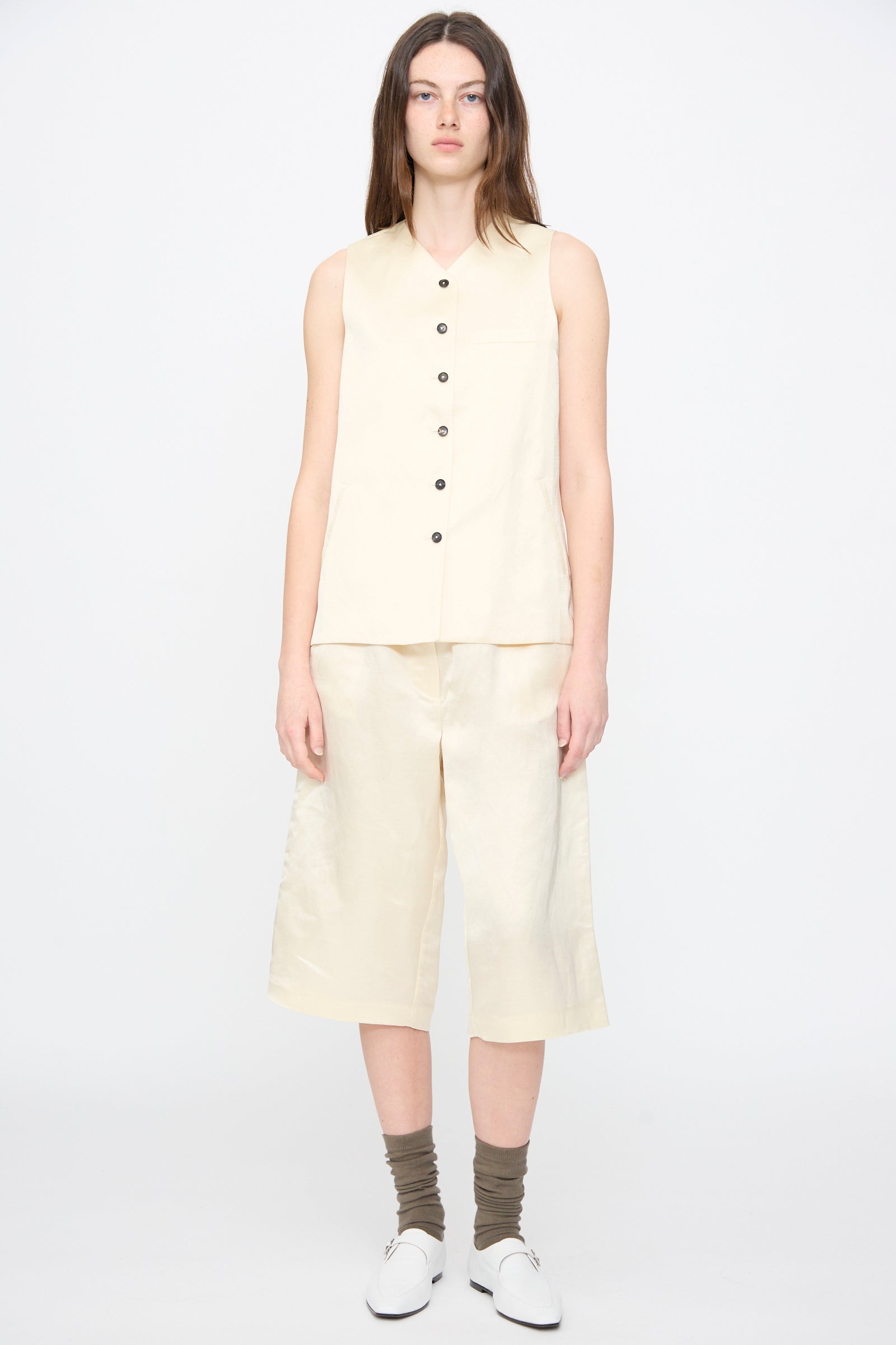 A person stands facing forward wearing the Satin Tailored Vest in Marfil by Cordera, matching knee-length shorts, gray socks, and white shoes against a plain light background.