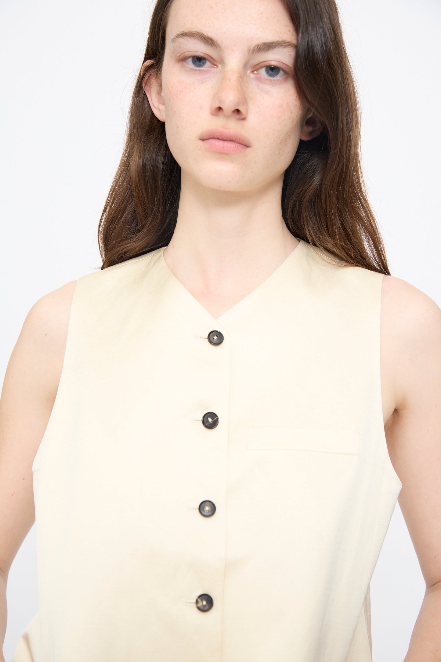 A woman with long brown hair wears the Cordera Satin Tailored Vest in Marfil, a sleeveless light beige piece with dark buttons, standing against a plain white background.