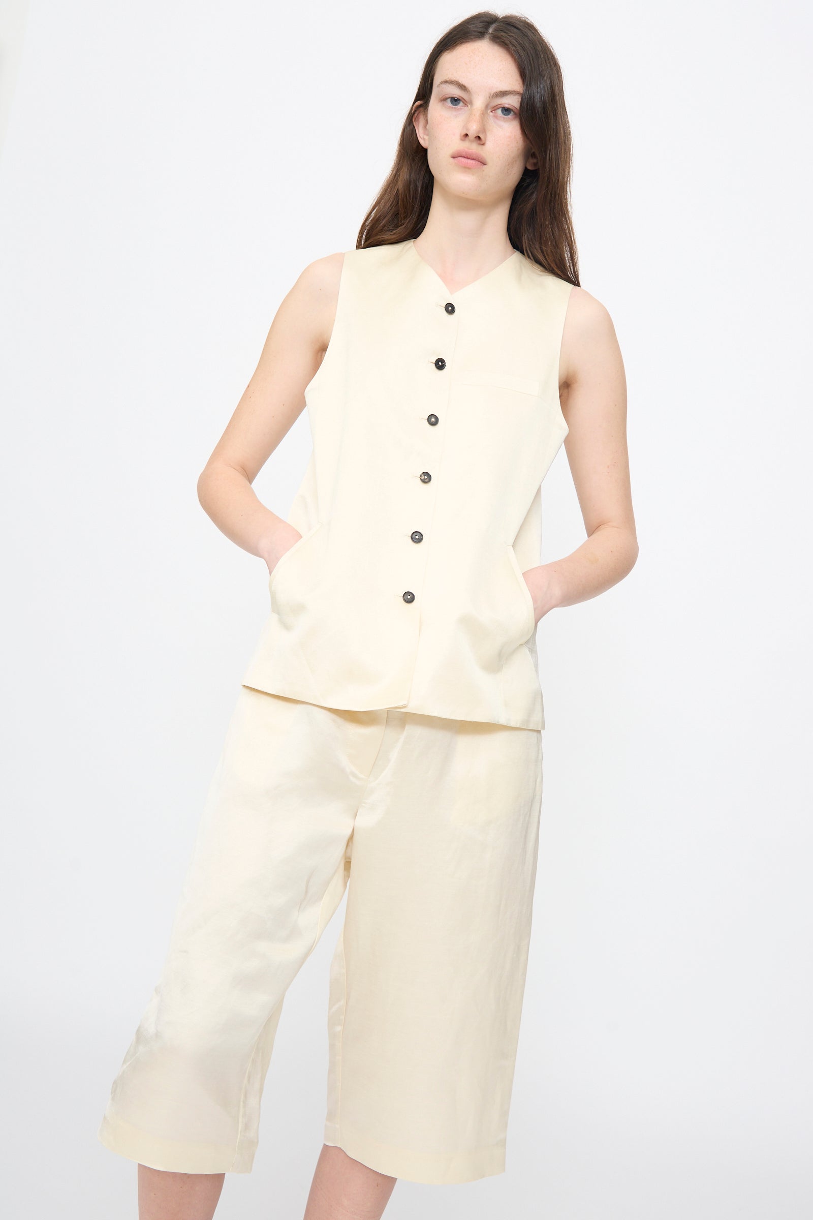 A person wears the Satin Tailored Vest in Marfil and matching knee-length shorts by Cordera, both made in Spain, standing hands-in-pockets against a plain background.