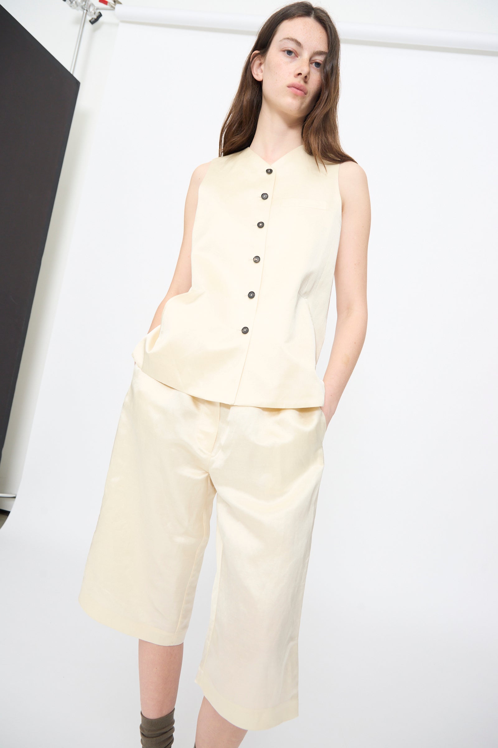 Someone models the Cordera Satin Tailored Vest in Marfil paired with matching knee-length shorts, standing with hands in pockets against a plain, light background.