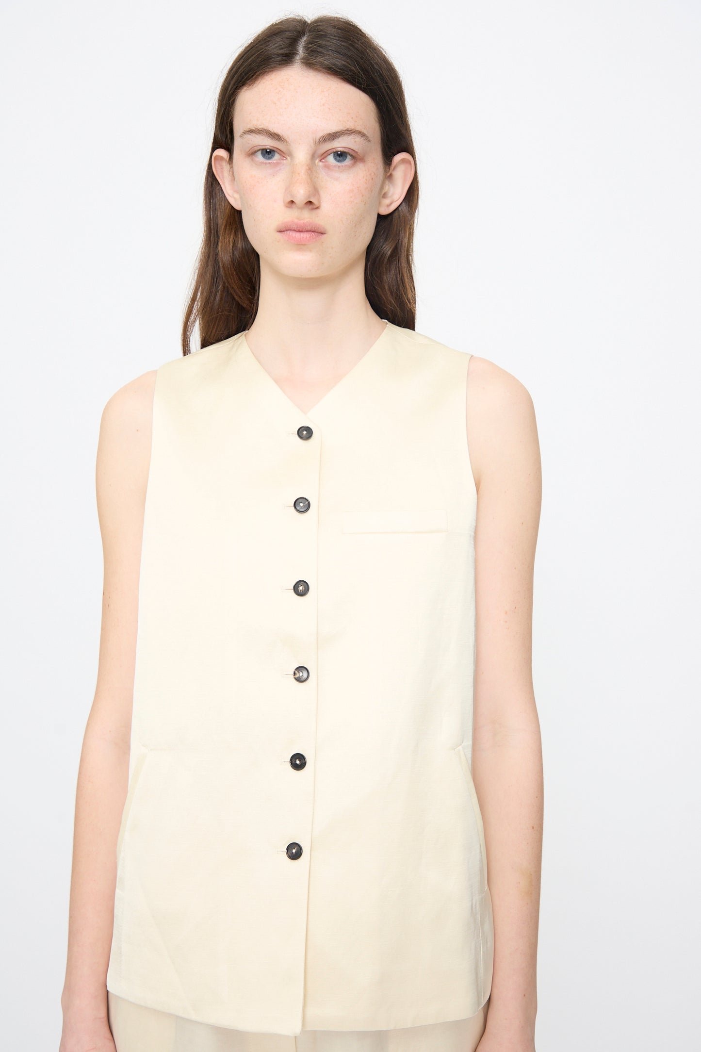 A woman with straight brown hair models the Cordera Satin Tailored Vest in Marfil, a sleeveless cream-colored piece with black buttons, against a plain white background.