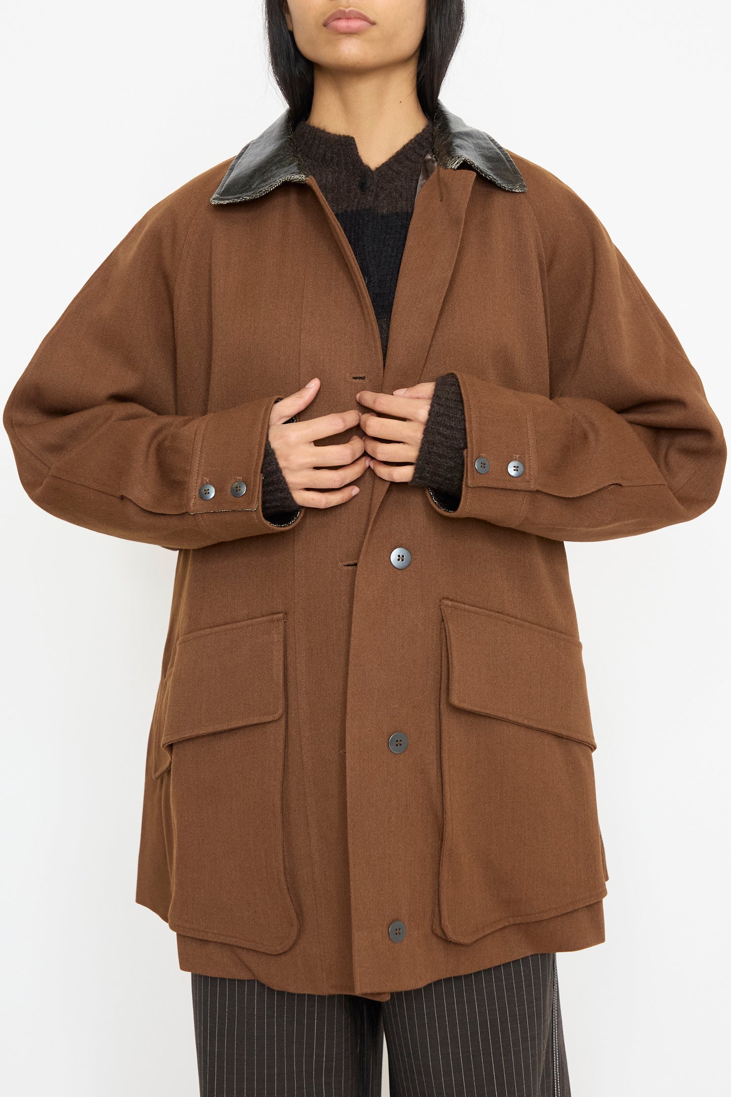 A person is wearing a Cordera Short Trench Coat in Root, featuring an oversized design with a dark collar and large pockets, crafted from virgin wool, over a black top, against a white backdrop. 