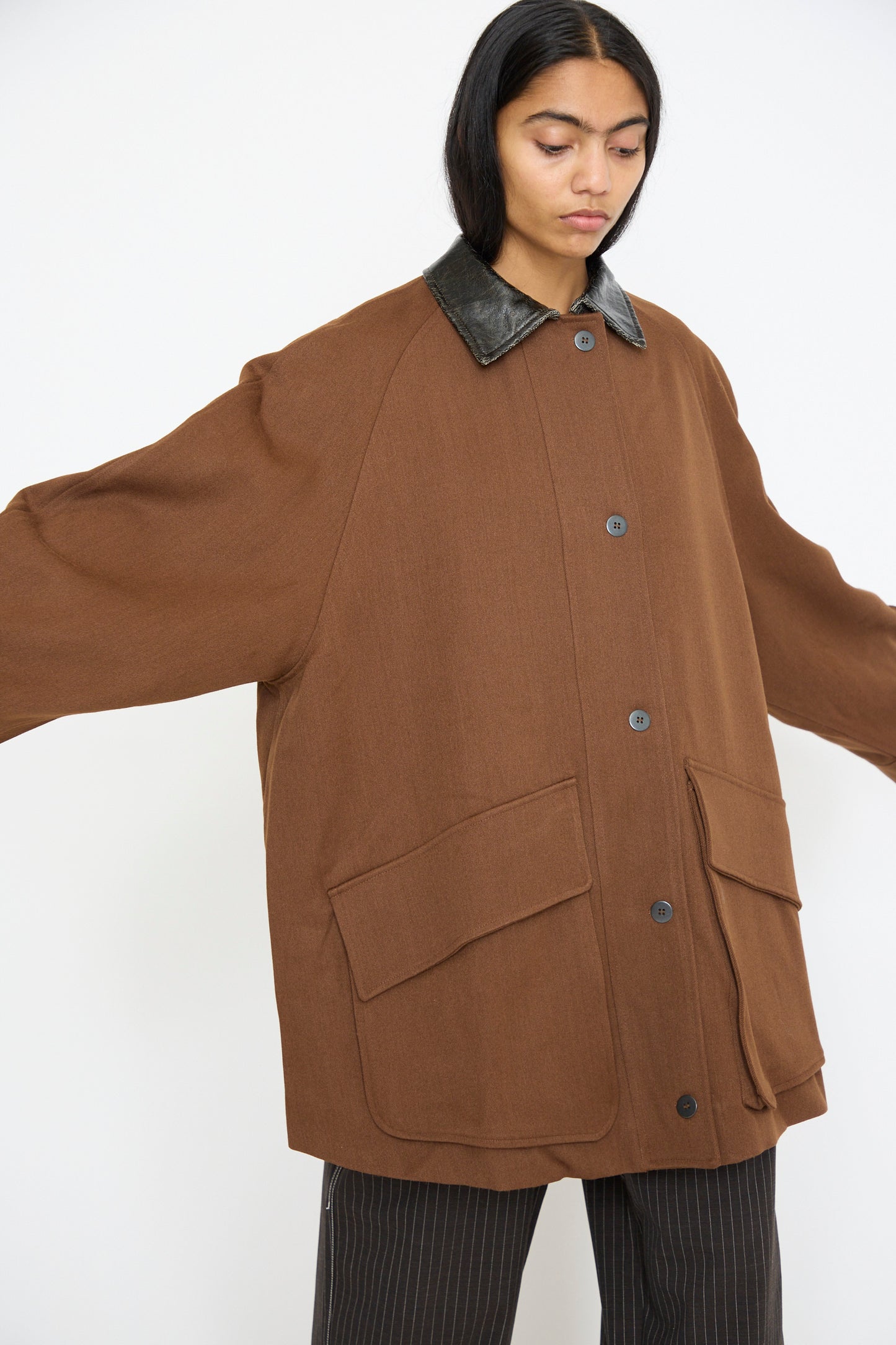 A person models the Cordera Short Trench Coat in Root, featuring an oversized fit with a black collar and large pockets. Arms are outstretched to display the design.