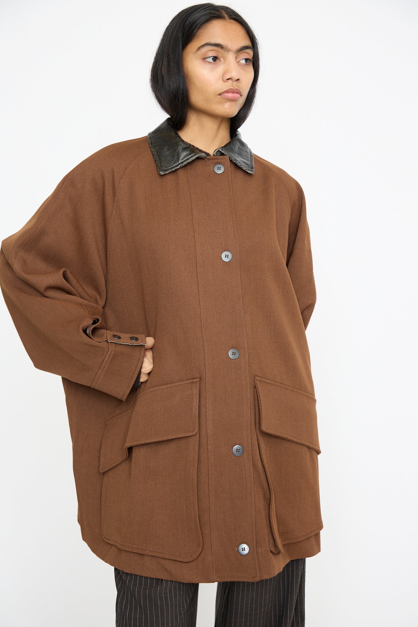 A person stands against a plain background, wrapped in an oversized Cordera Short Trench Coat in Root, featuring a black collar and buttons, crafted from luxurious virgin wool.