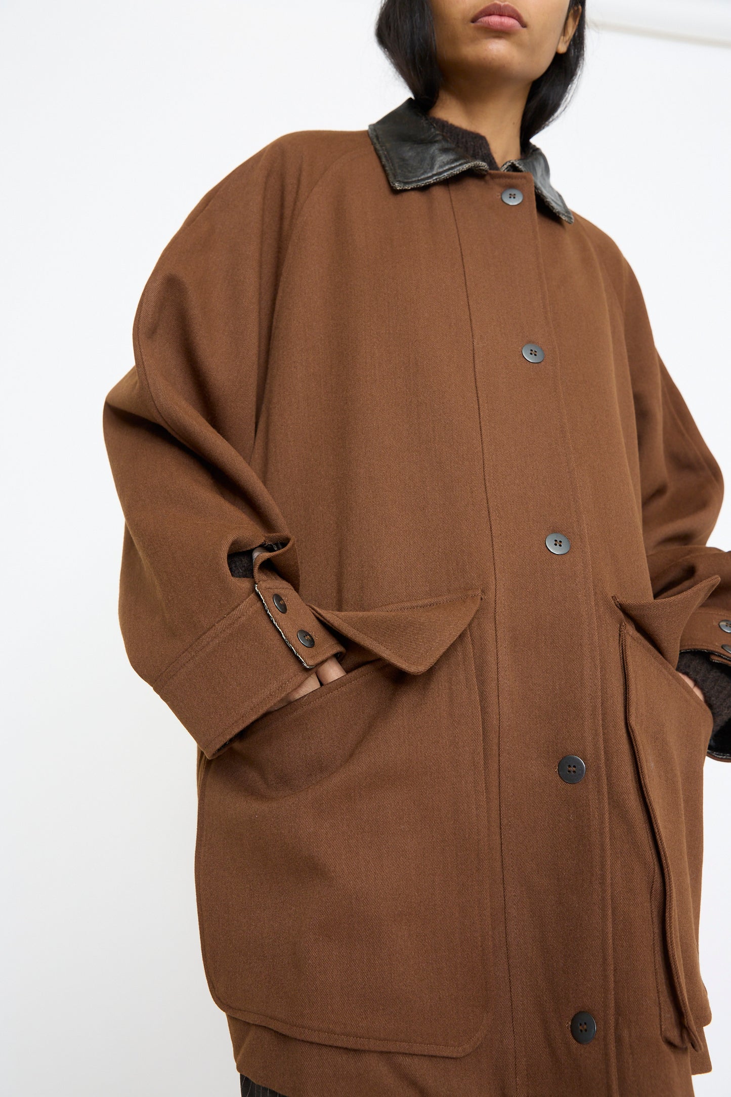 A person wears Cordera's Short Trench Coat in Root, an oversized design made from virgin wool, featuring a black leather collar and large front pockets.