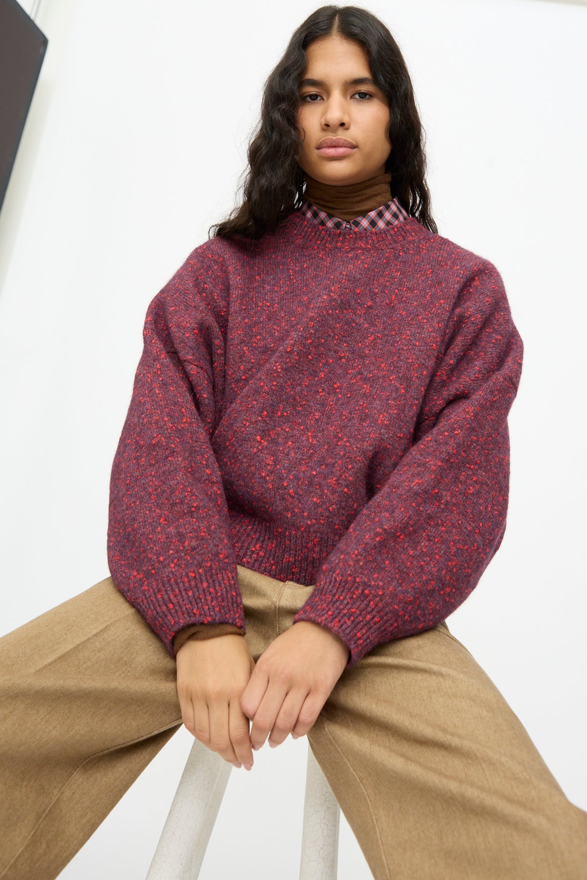 A person wearing a Cordera Speckled Crewneck Sweater in Beaujolais over a collared shirt, paired with tan trousers, sitting on a stool.