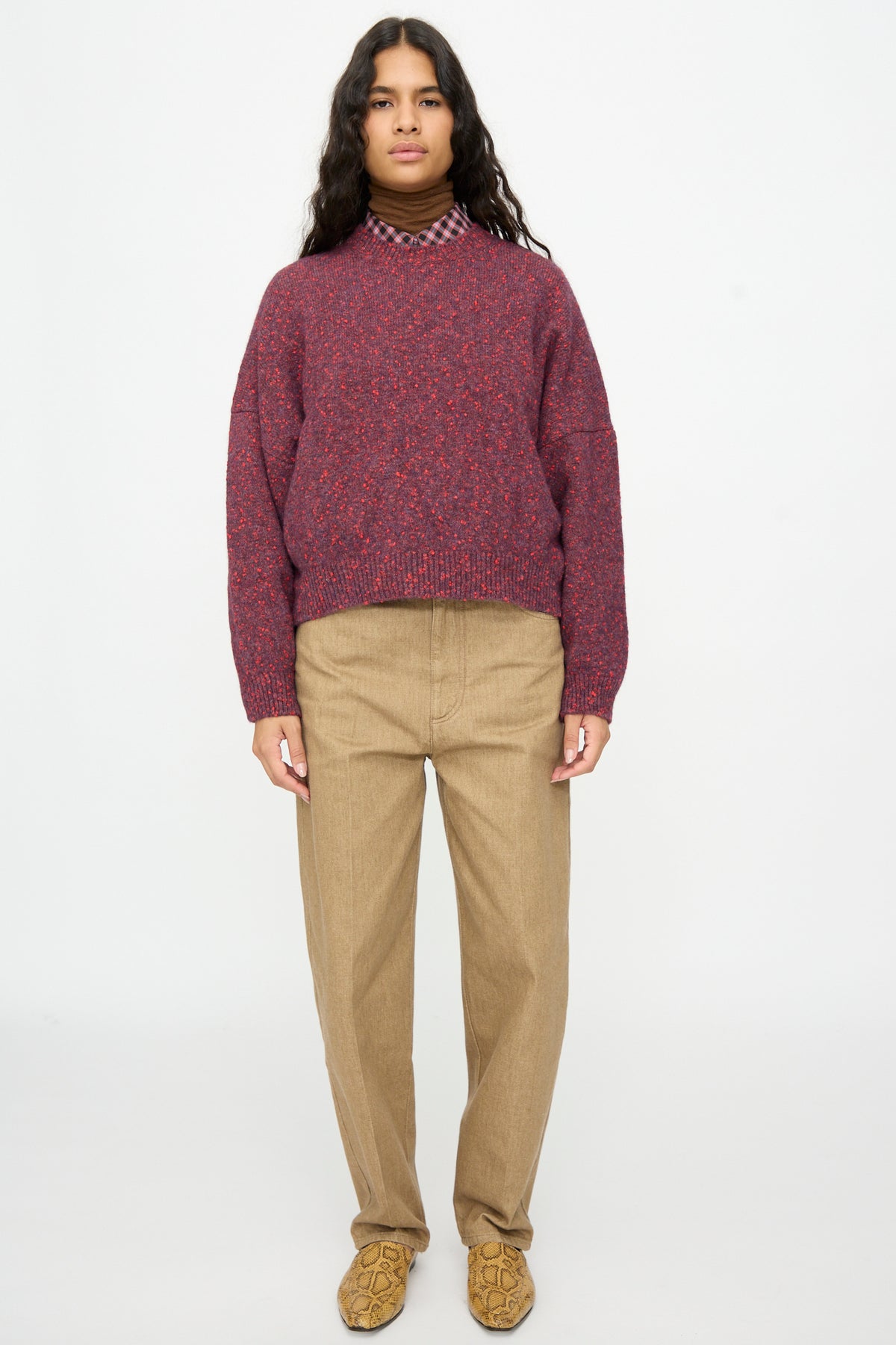 A person wears a boxy-fit Cordera Speckled Crewneck Sweater in Beaujolais layered over a brown turtleneck and plaid shirt, paired with tan trousers and snakeskin-print shoes.