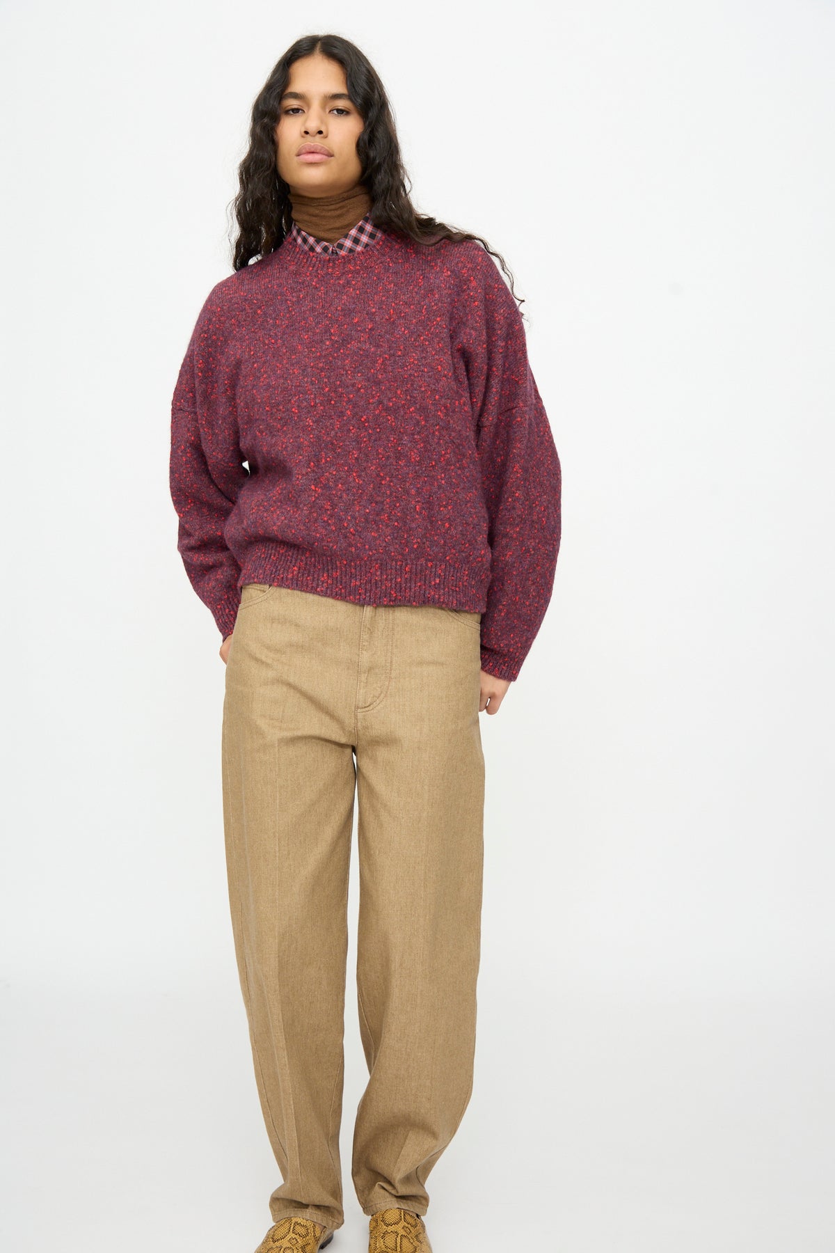 A person wears a Cordera Speckled Crewneck Sweater in Beaujolais over a brown turtleneck and plaid shirt, paired with tan trousers.
