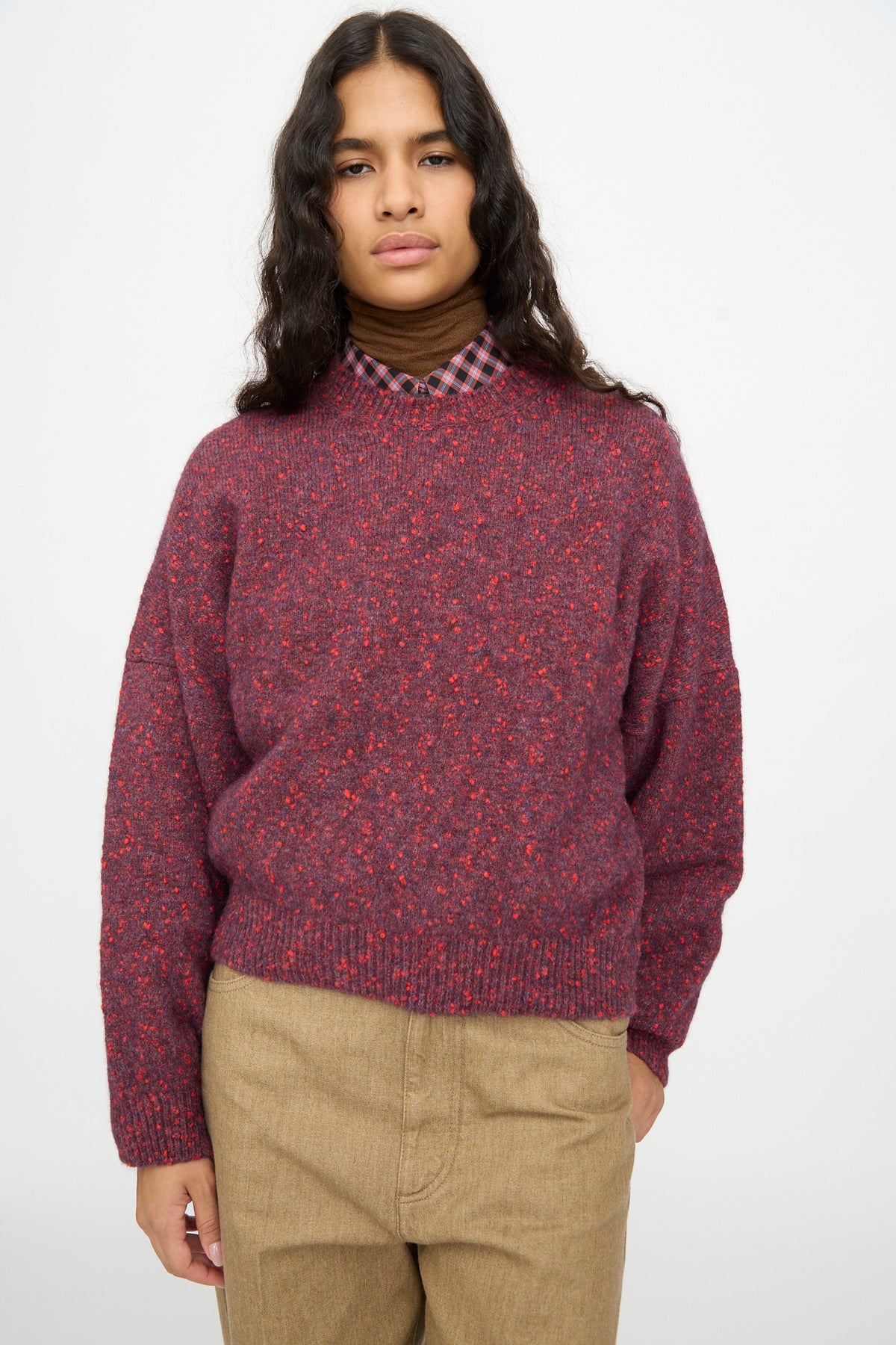 A person wears a Cordera Speckled Crewneck Sweater in Beaujolais layered over a plaid collared shirt and brown trousers.