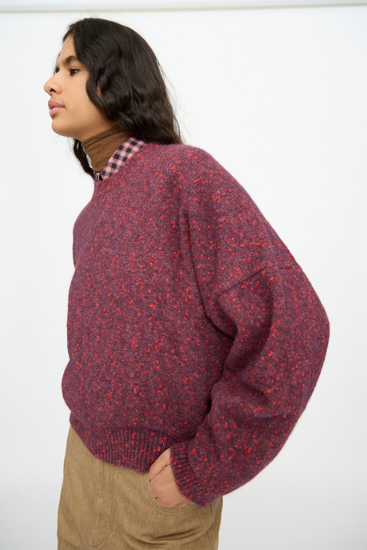 A person wears a Cordera Speckled Crewneck Sweater in Beaujolais over a plaid collared shirt and tan trousers.