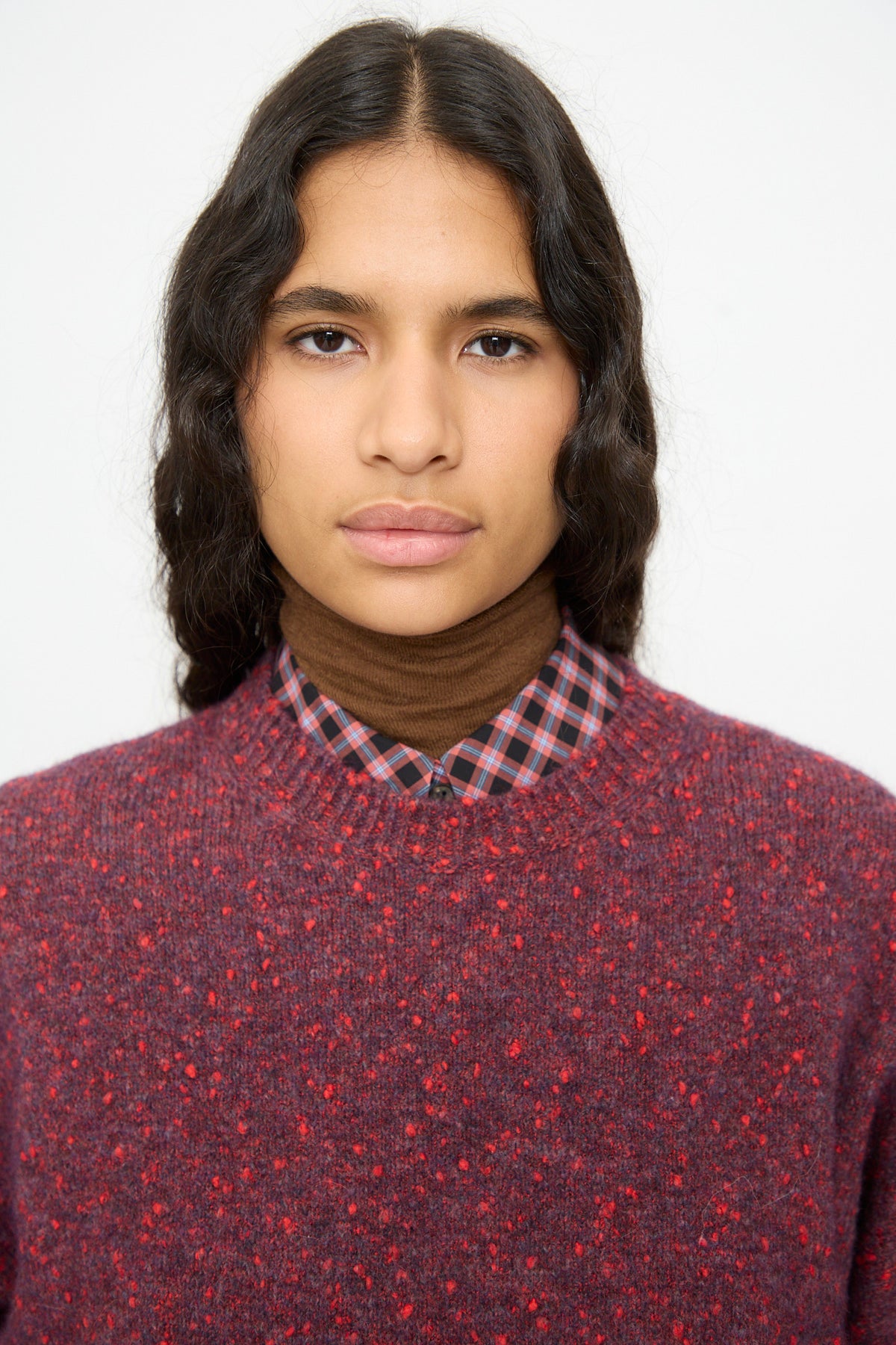 A person wearing a Cordera Speckled Crewneck Sweater in Beaujolais, a relaxed-fit, maroon speckled crewneck layered over a plaid shirt and a brown turtleneck.