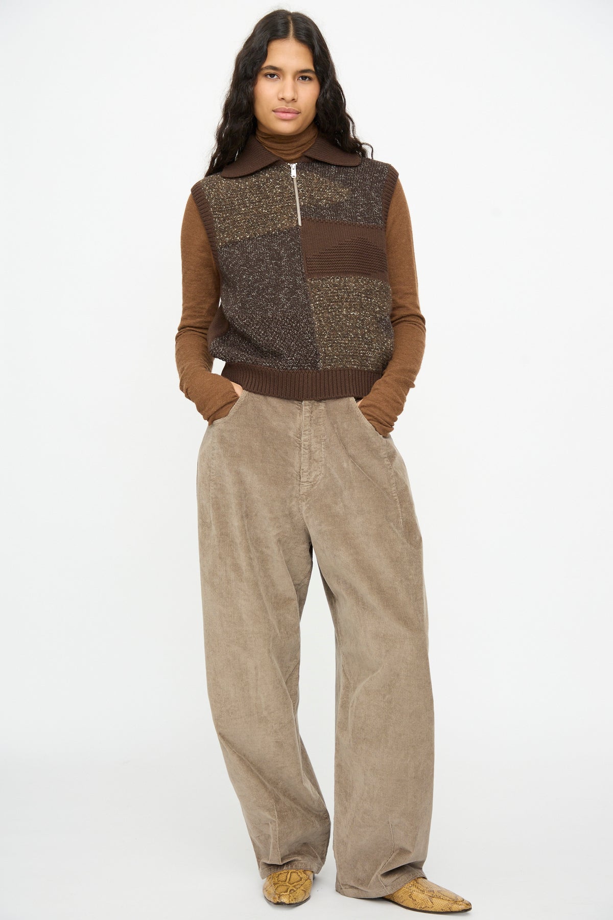 A person wearing a Cordera Speckled Waistcoat in Brown over a long-sleeved turtleneck, paired with wide-leg beige trousers and patterned pointed-toe shoes.