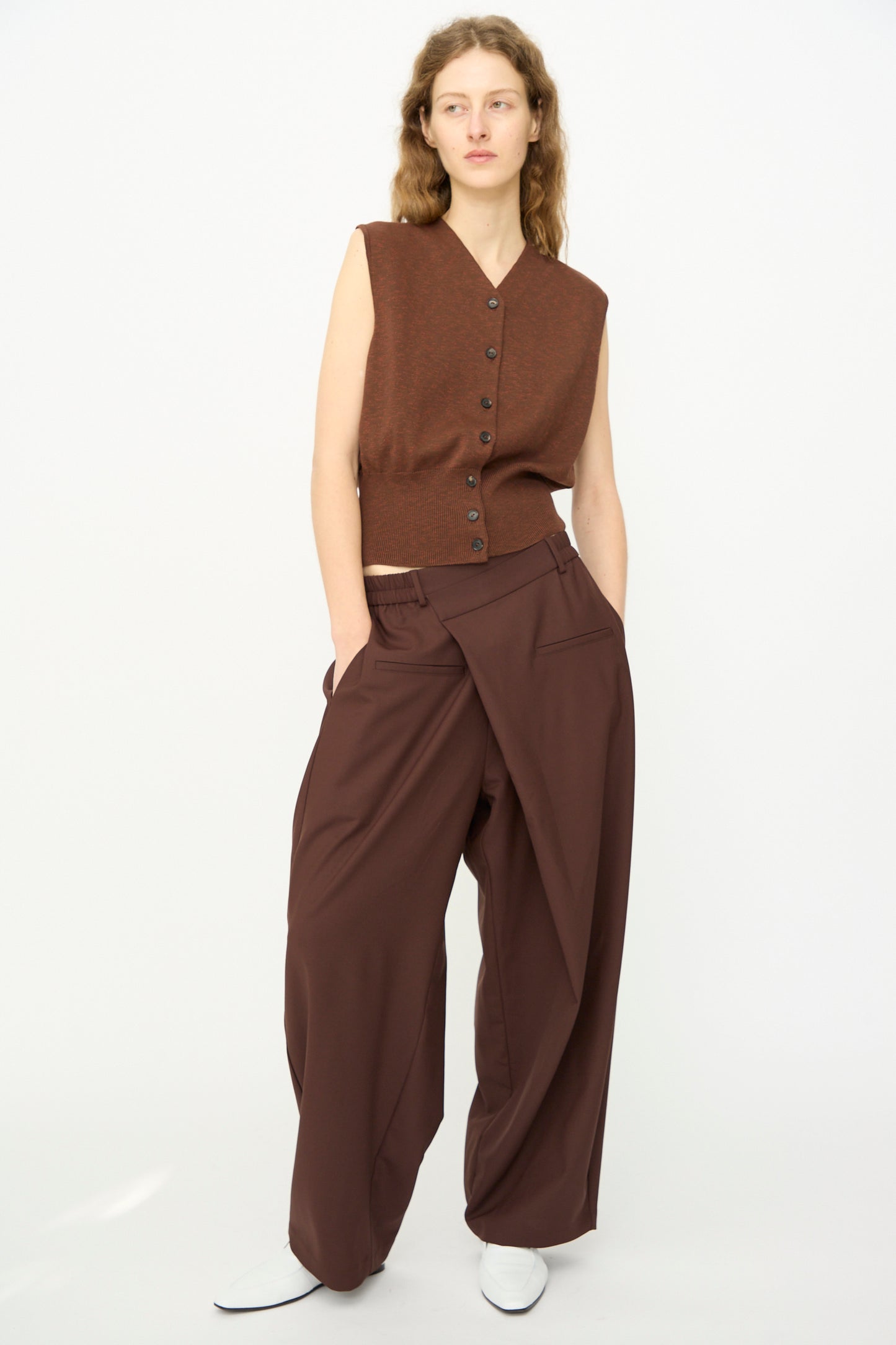 A person stands against a plain background wearing Cordera’s Tailored Crossover Pant in Burgundy with a sleeveless brown buttoned top and white shoes, hands in pockets.