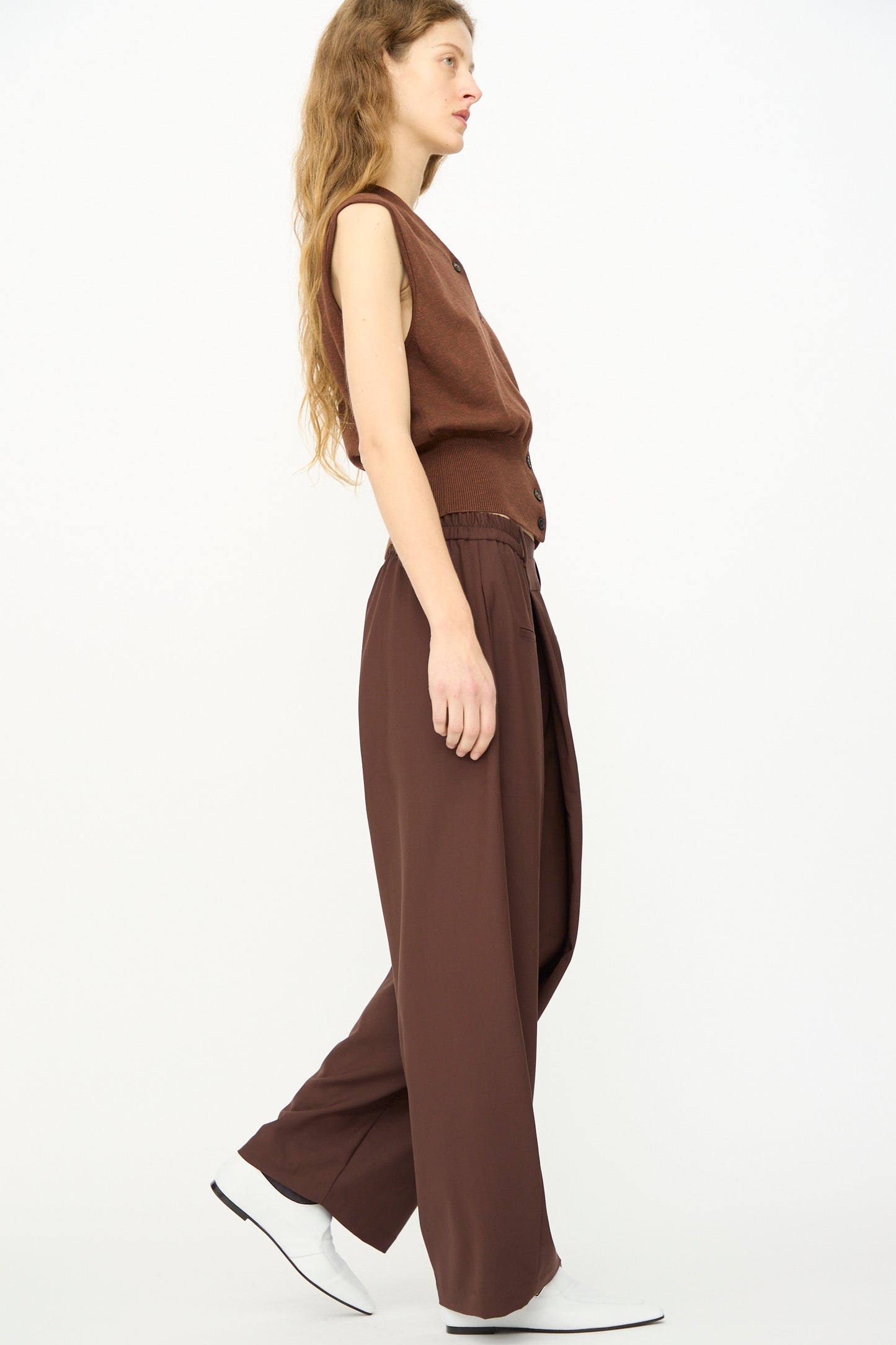 Person with long hair in a sleeveless brown top and Cordera’s Tailored Crossover Pant in Burgundy, walking to the left against a plain white background.