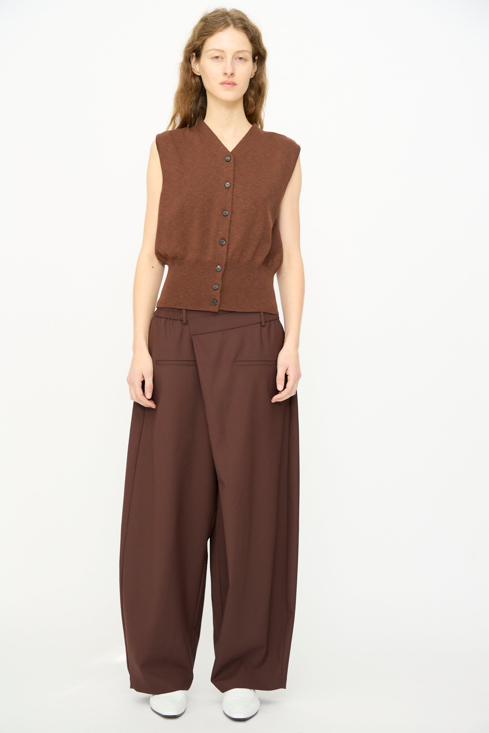 A woman stands against a plain background wearing a sleeveless brown button-up top, Cordera’s Tailored Crossover Pant in Burgundy, and white shoes.