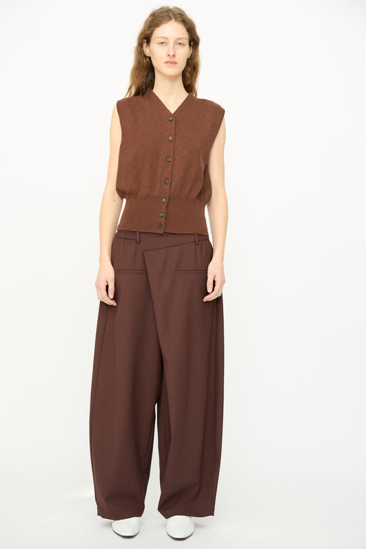 A woman stands against a plain background wearing a sleeveless brown button-up top, Cordera’s Tailored Crossover Pant in Burgundy, and white shoes.
