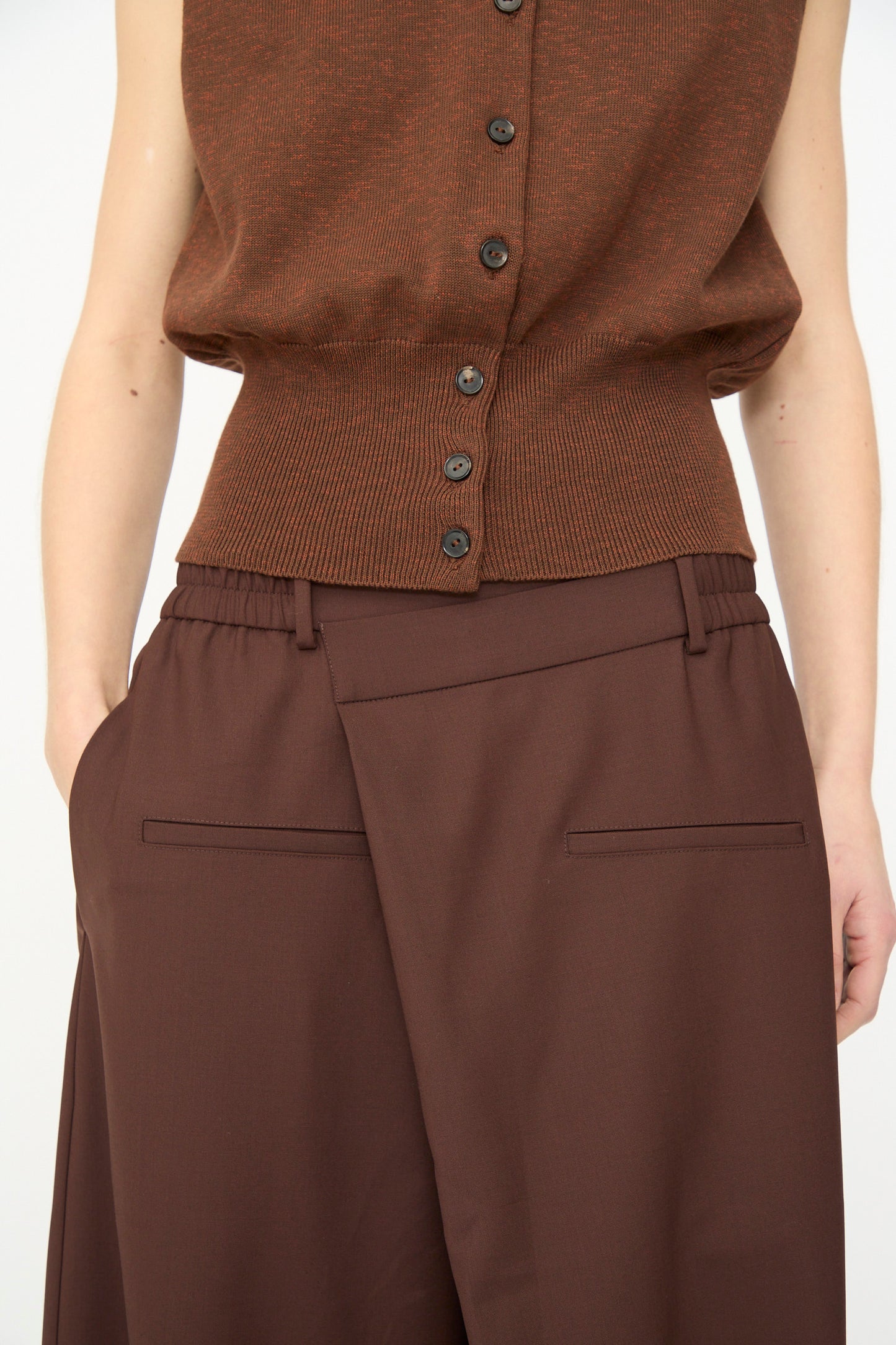 A person wears a brown sleeveless button-up top with Cordera's Tailored Crossover Pant in Burgundy, styled high-waisted and wide-leg, one hand in a pocket, shown from shoulders to thighs.