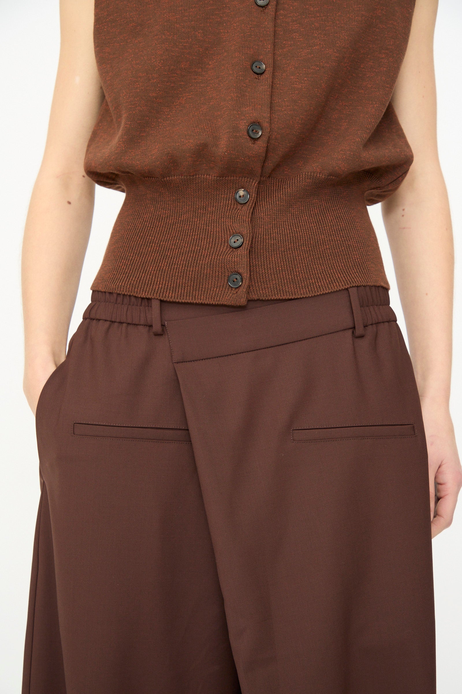 A person wears a brown sleeveless button-up top with Cordera's Tailored Crossover Pant in Burgundy, styled high-waisted and wide-leg, one hand in a pocket, shown from shoulders to thighs.