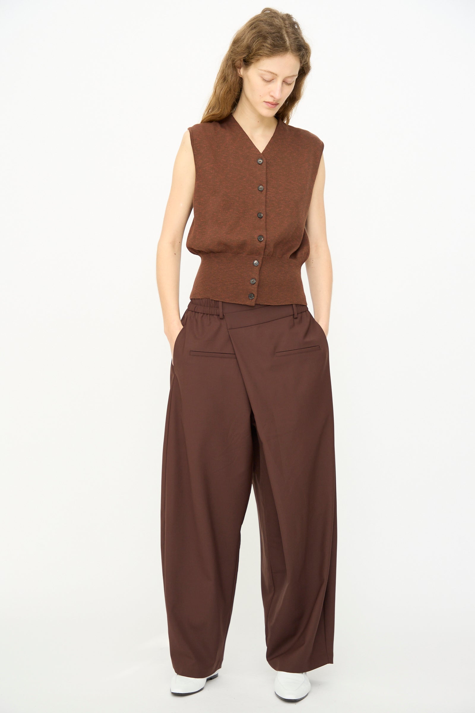 A person wearing a sleeveless brown buttoned vest and Cordera’s Tailored Crossover Pant in Burgundy, with hands in pockets, stands against a plain white background.