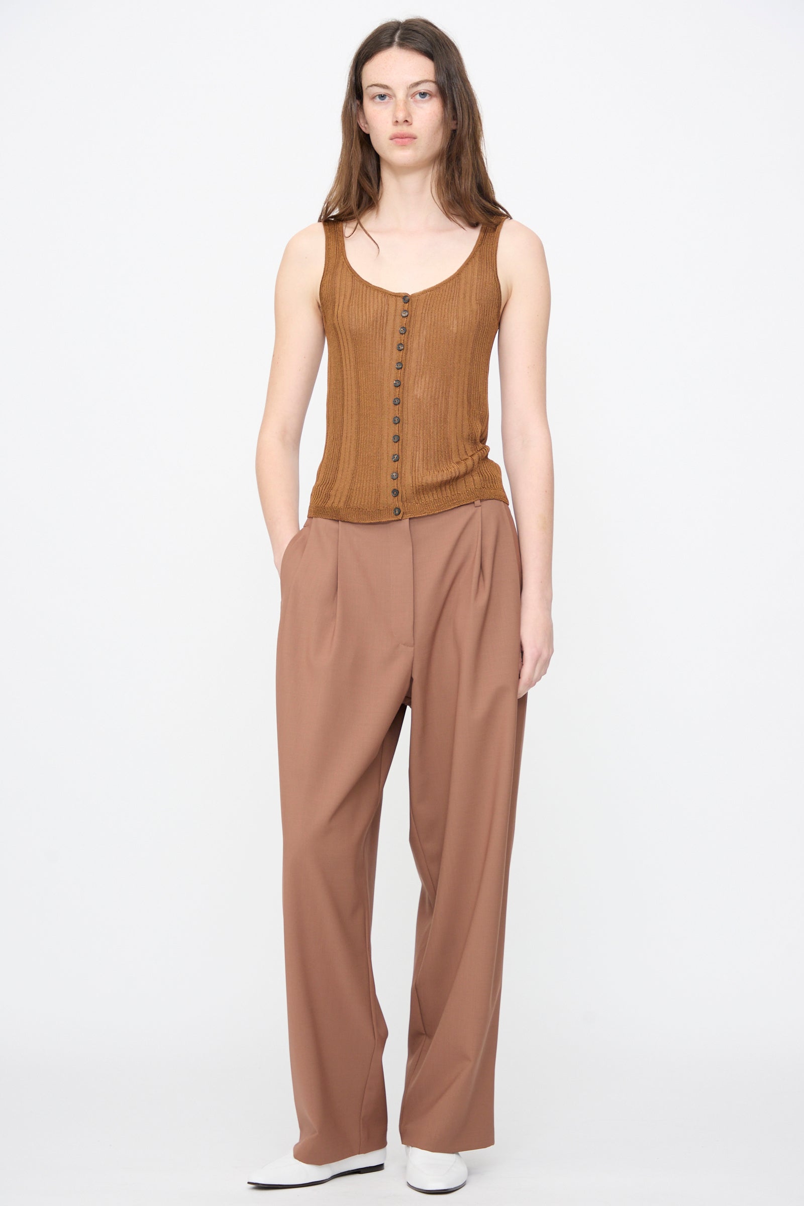 A person stands against a plain white background wearing Cordera's Tailored Masculine Pant in Clove, paired with a brown sleeveless buttoned top and white shoes.