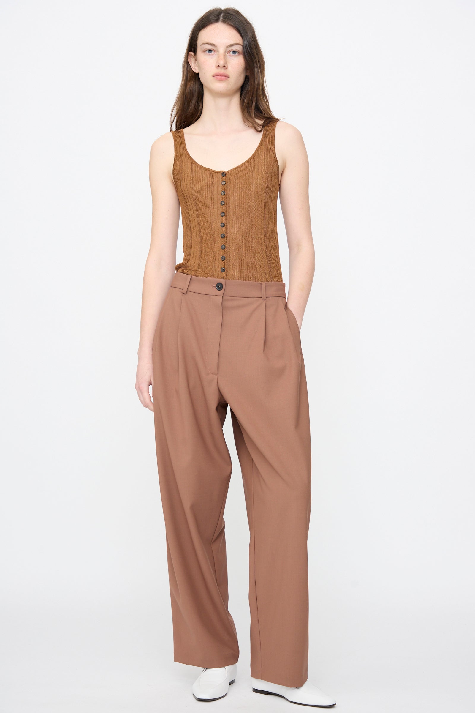 A person stands against a plain background wearing a sleeveless brown ribbed top, Cordera's Tailored Masculine Pant in Clove with a high-waisted relaxed fit, and white shoes, hands in pockets.