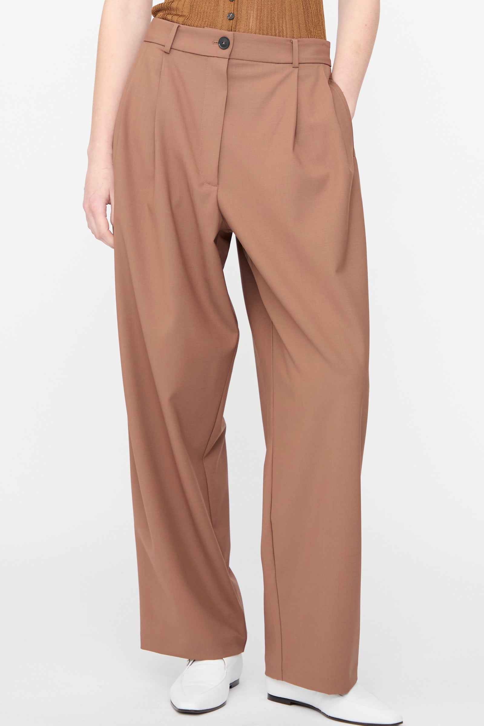 Model wearing Cordera's Tailored Masculine Pant in Clove, a high-waisted, pleated relaxed fit, paired with a tucked-in brown top and white shoes against a plain light background.