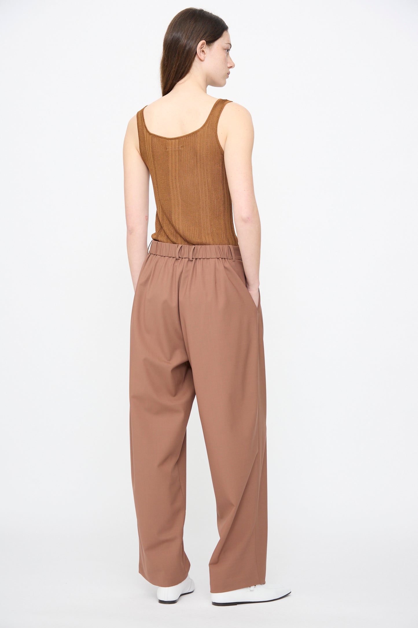 A person faces away against a white background, wearing Cordera’s Tailored Masculine Pant in Clove with a brown sleeveless top and white shoes.