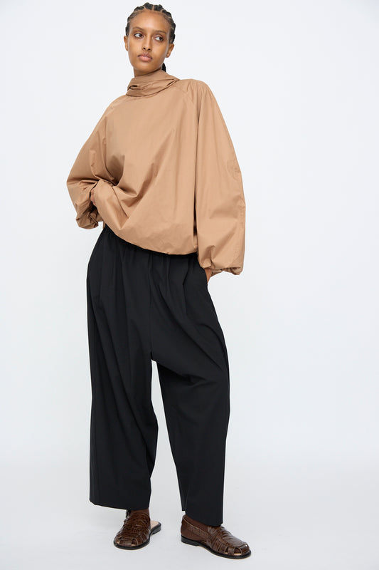 Person standing against a plain background, wearing a loose tan long-sleeve top, Cordera Tailoring Maxi Pants in Black with an elastic waistband, and brown shoes.