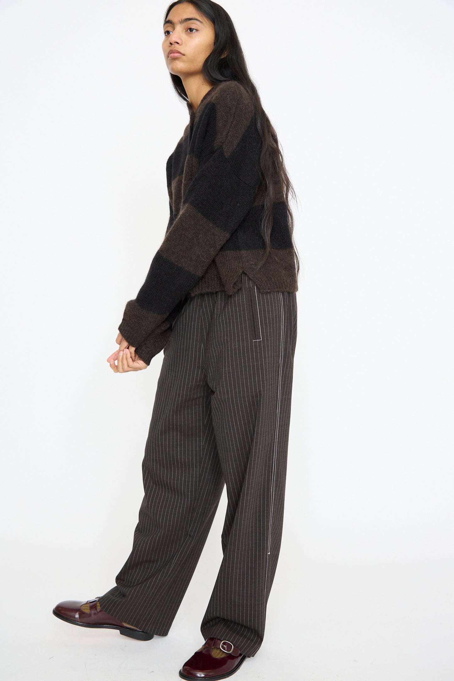 A person stands against a white background wearing a brown and black striped sweater with Cordera's Tailoring Relaxed Pant in Shade, featuring pinstripes and a drawstring waist. 